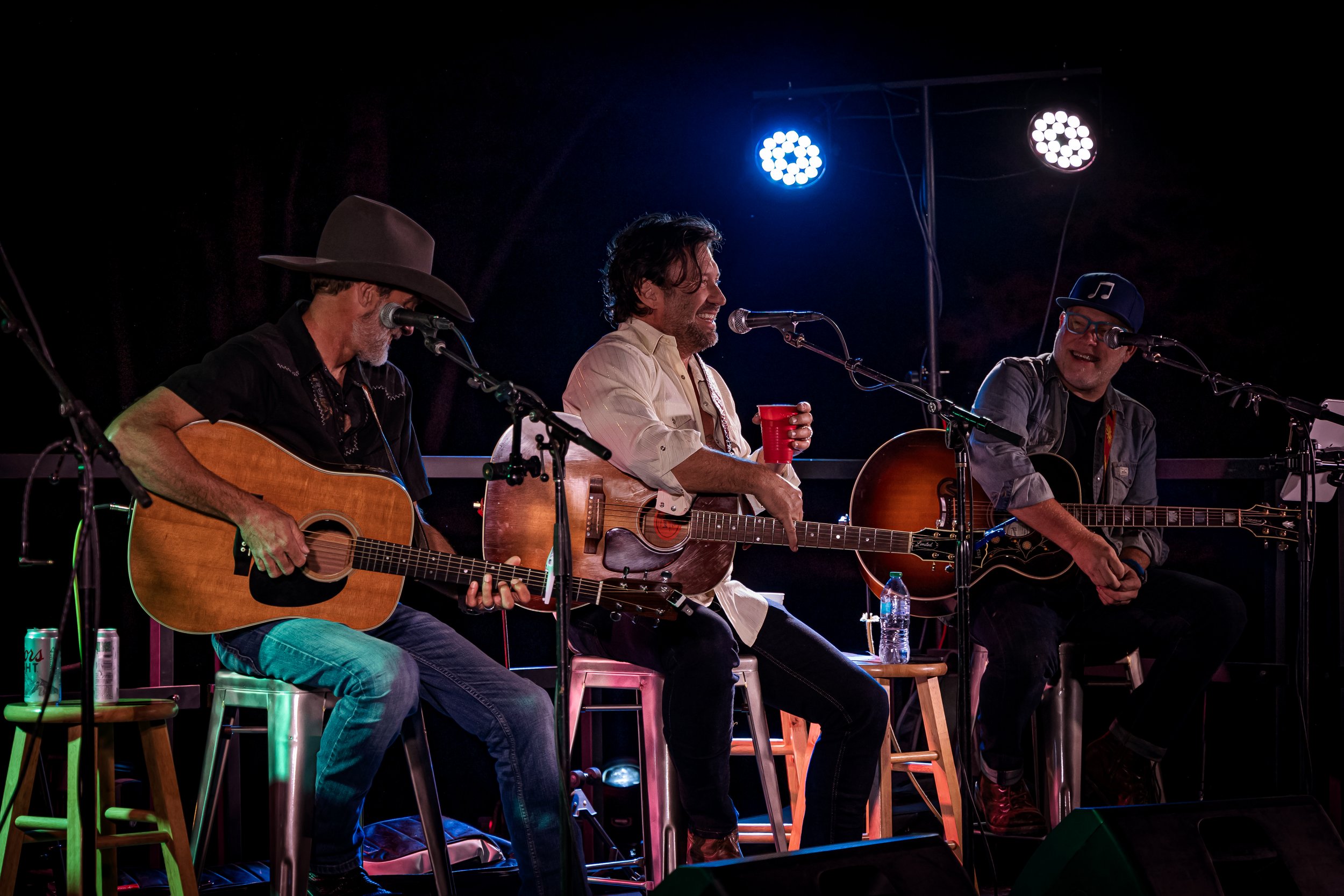 Three men playing acoustic guitars on stage during a live performance, with colorful stage lighting, two are singing into microphones, one is holding a red cup, and water bottles are on small stools beside them.