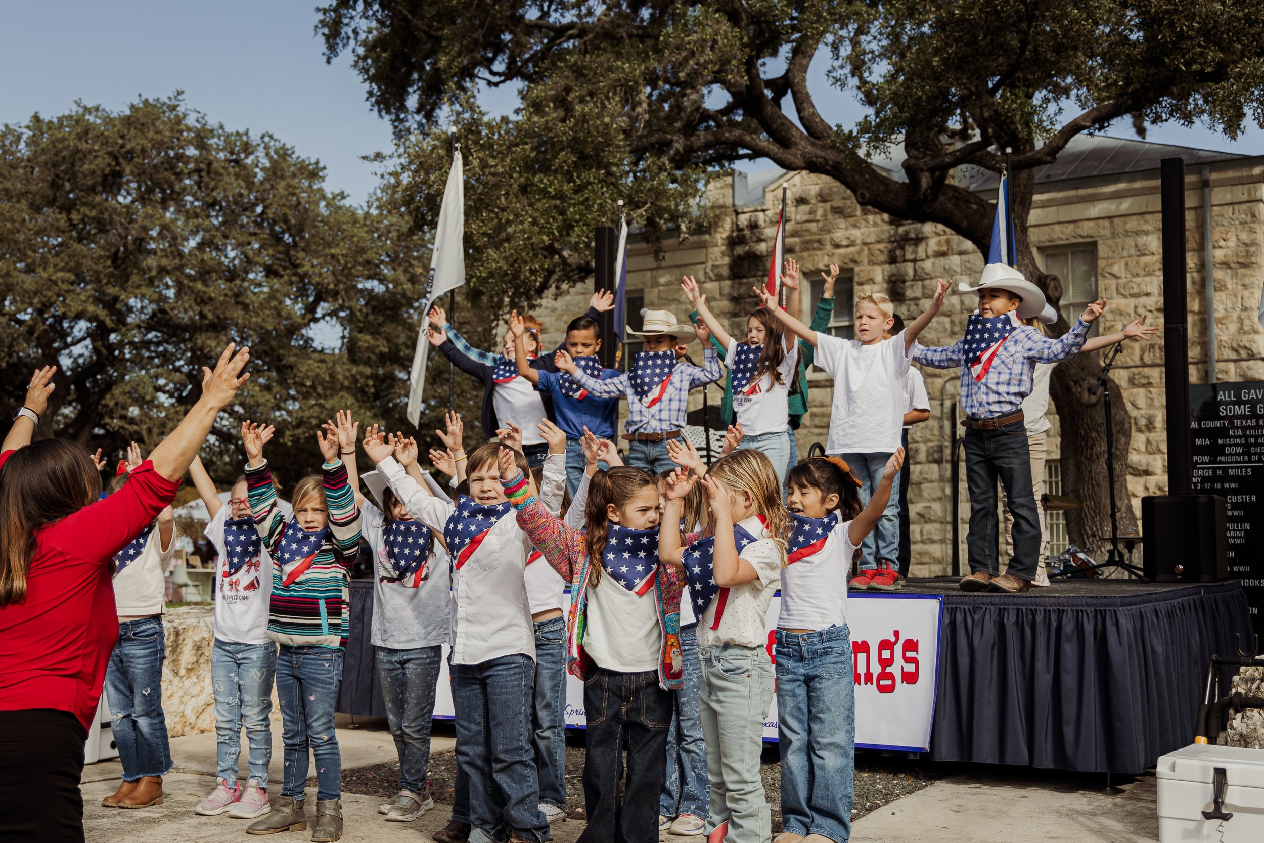 Children on a stage wearing patriotic bandanas and hats performing during a celebration, with an adult leading them in front, and trees and flags in the background.