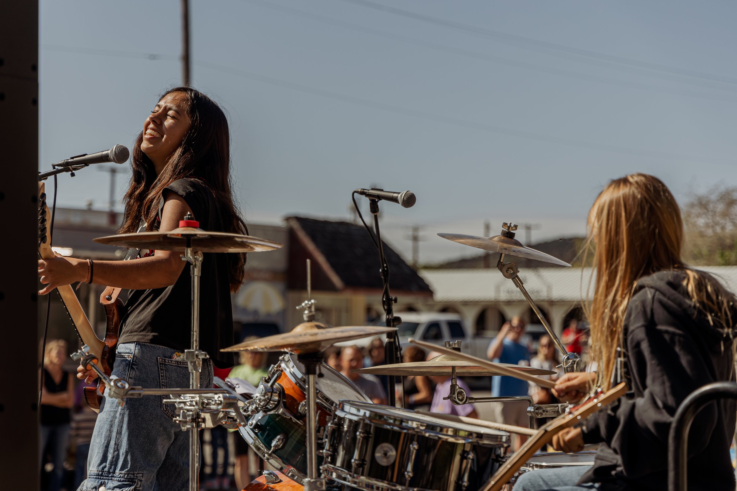 Two young women performing music outdoors, one singing and playing guitar, the other playing drums, with an audience in the background.