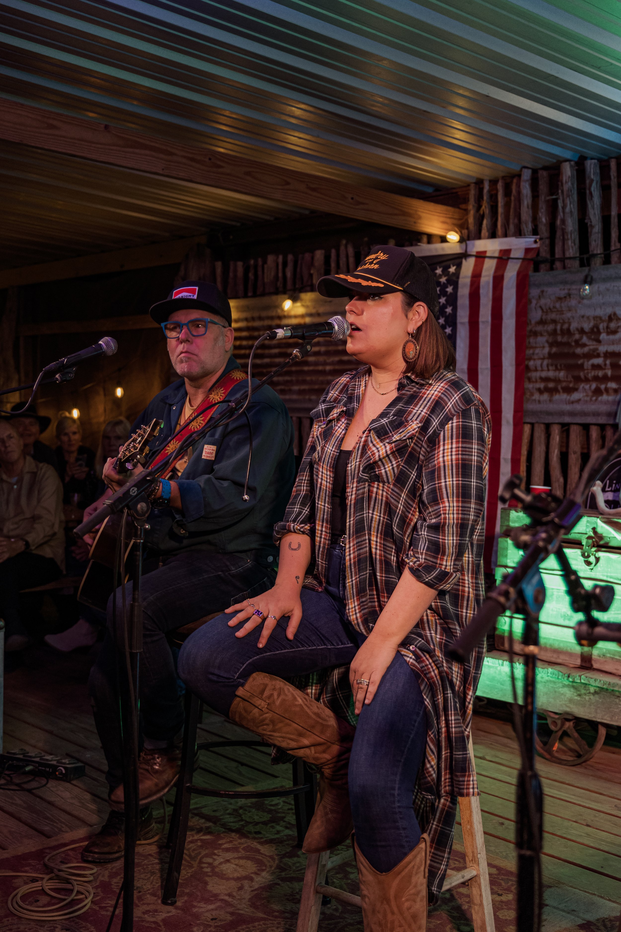 A woman and a man singing and playing guitar during a live performance in a rustic venue with an American flag in the background.