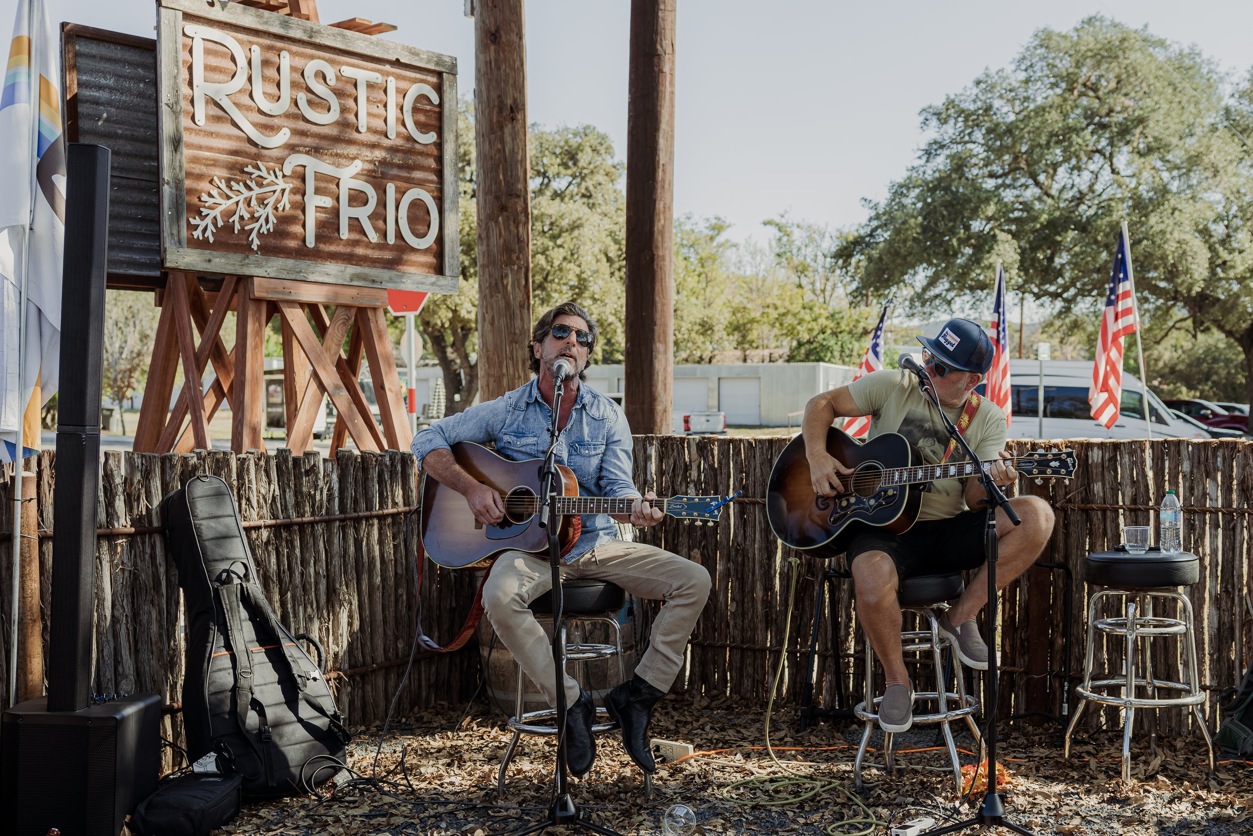 Two men playing acoustic guitars and singing at an outdoor event with a Rusted Rio sign, American flags, and trees in the background.