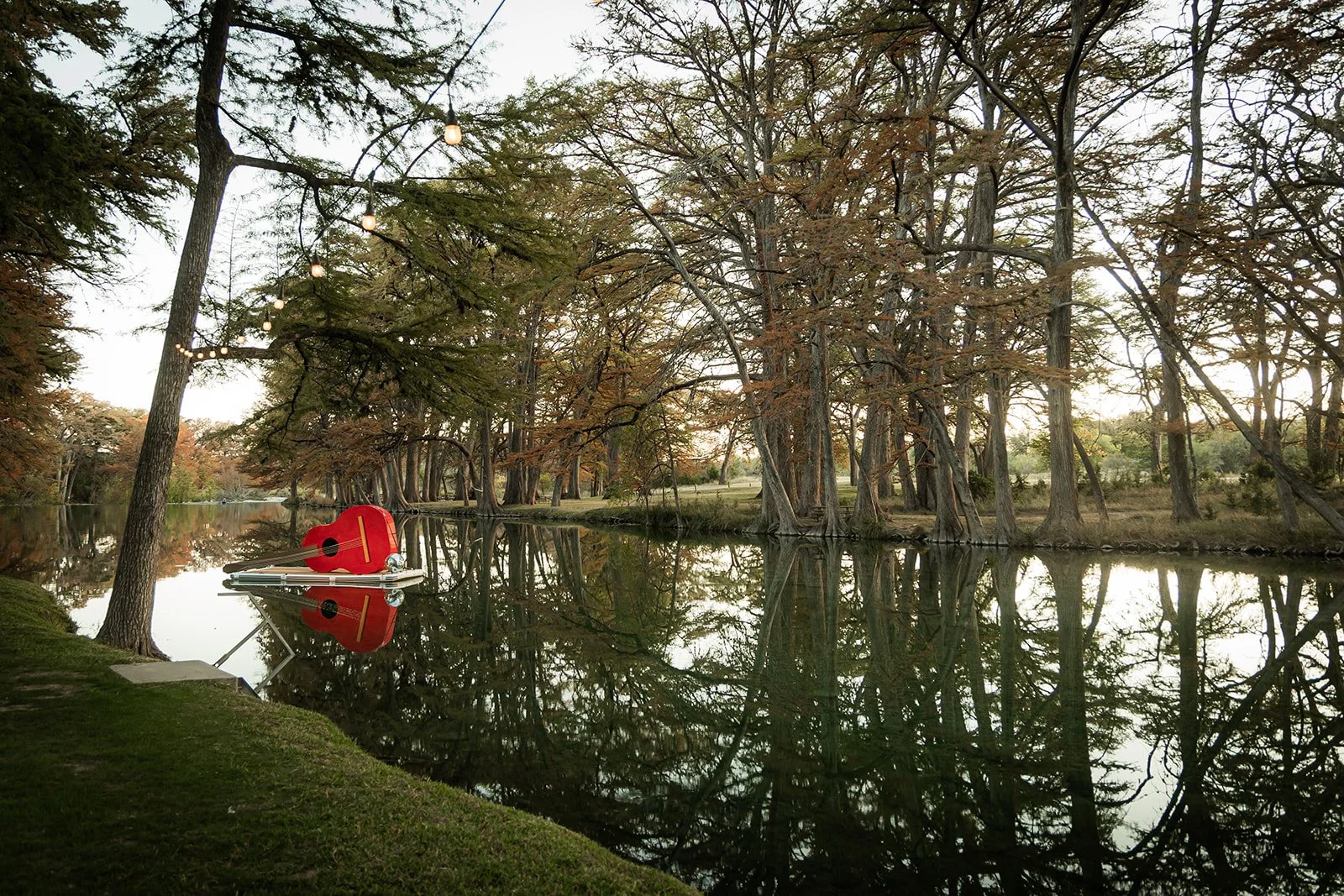 A small boat with a red guitar-shaped cover floats on a calm river, surrounded by trees with autumn foliage and string lights hanging overhead.