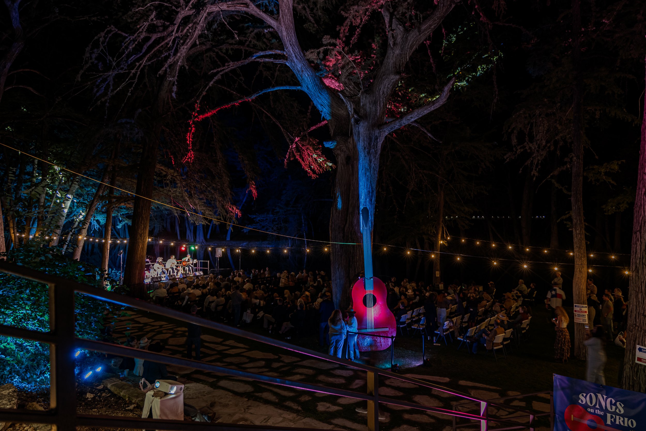 A nighttime outdoor concert in a wooded area with a stage, a band, and audience seated in chairs. A large tree in the center is decorated with neon lights, including a giant illuminated guitar sculpture and painted blue and red accents. String lights