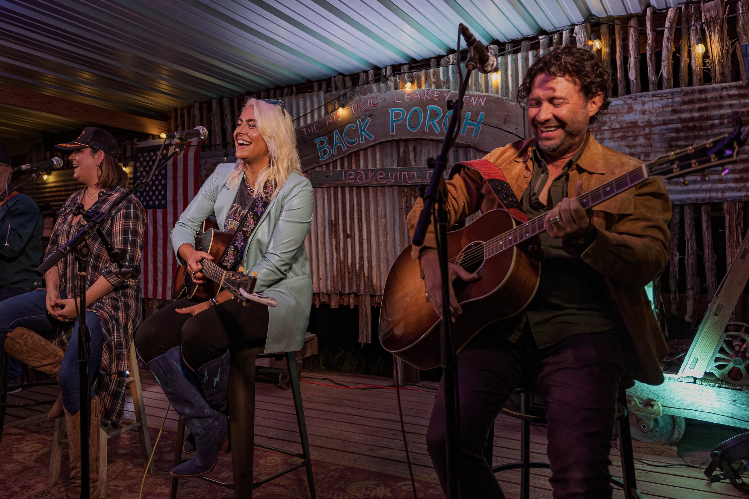 Musicians performing on stage at Back Porch venue, including a woman with blonde hair playing guitar and a man with curly hair playing guitar and singing, with rustic decor and American flag in background.