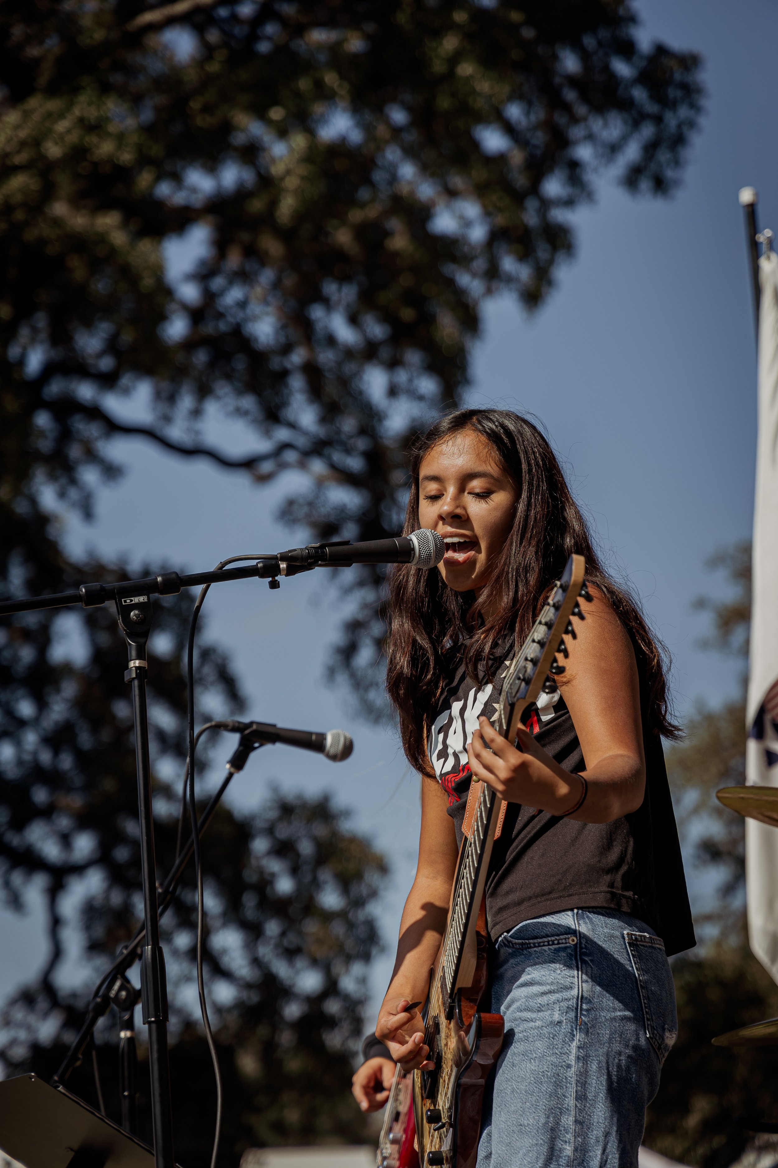 A young woman singing and playing an electric guitar outdoors during the daytime.