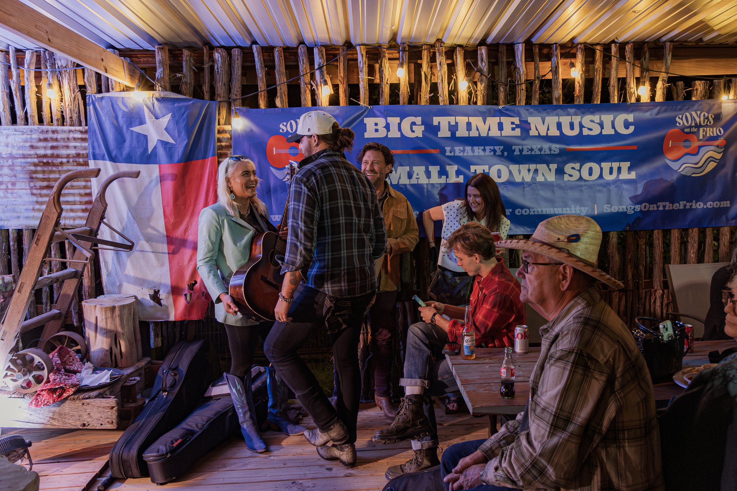 People gather in a rustic outdoor setting with a stage, a Texas flag, and a blue banner reading 'Big Time Music' and 'Small Town Soul,' as musicians perform and audience members enjoy the scene.