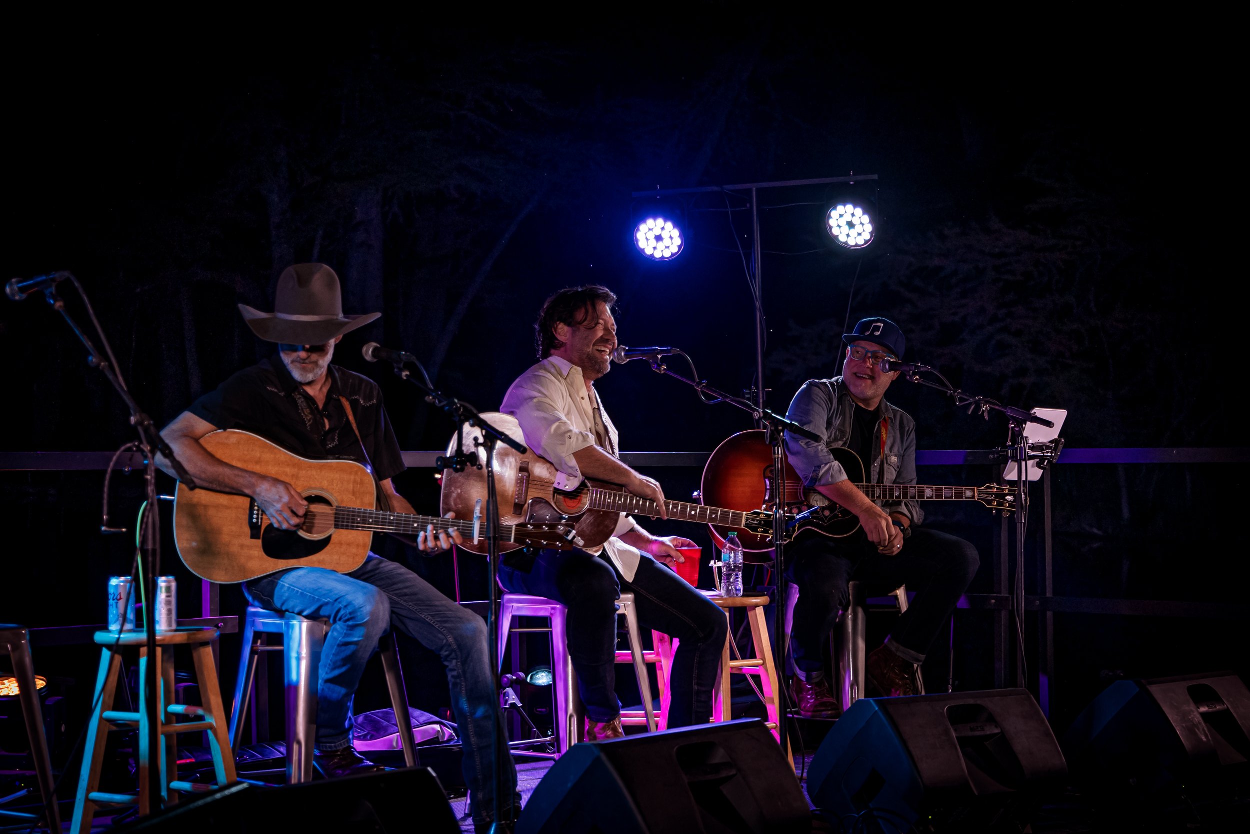Three male musicians performing on an outdoor stage at night, playing acoustic guitars and singing. They are illuminated by stage lights with dark trees in the background.