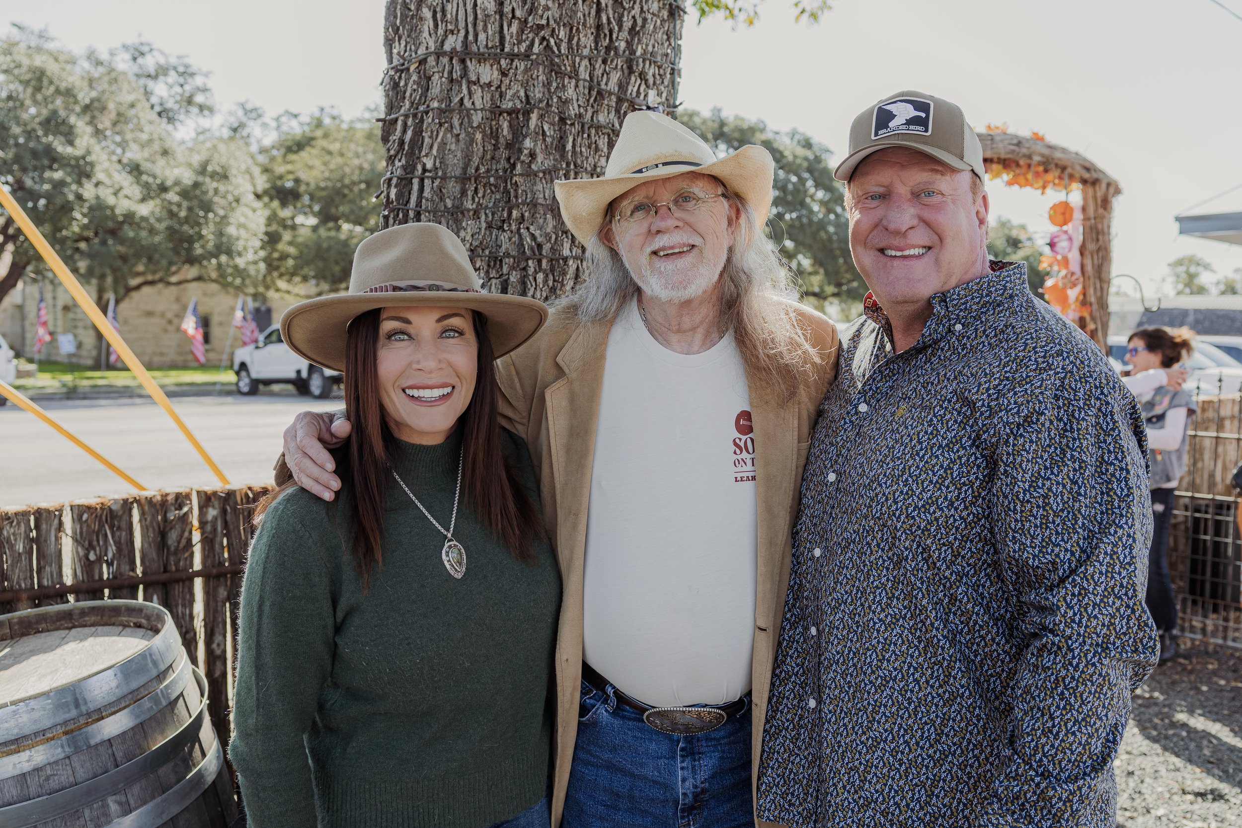 Group of three people smiling outdoors near a large tree, with a fence and onlookers in the background.