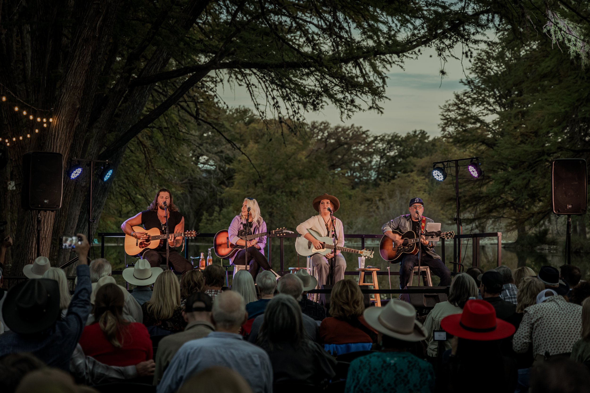 A band of four musicians playing guitars and singing on an outdoor stage during sunset, with an audience watching under trees and string lights.