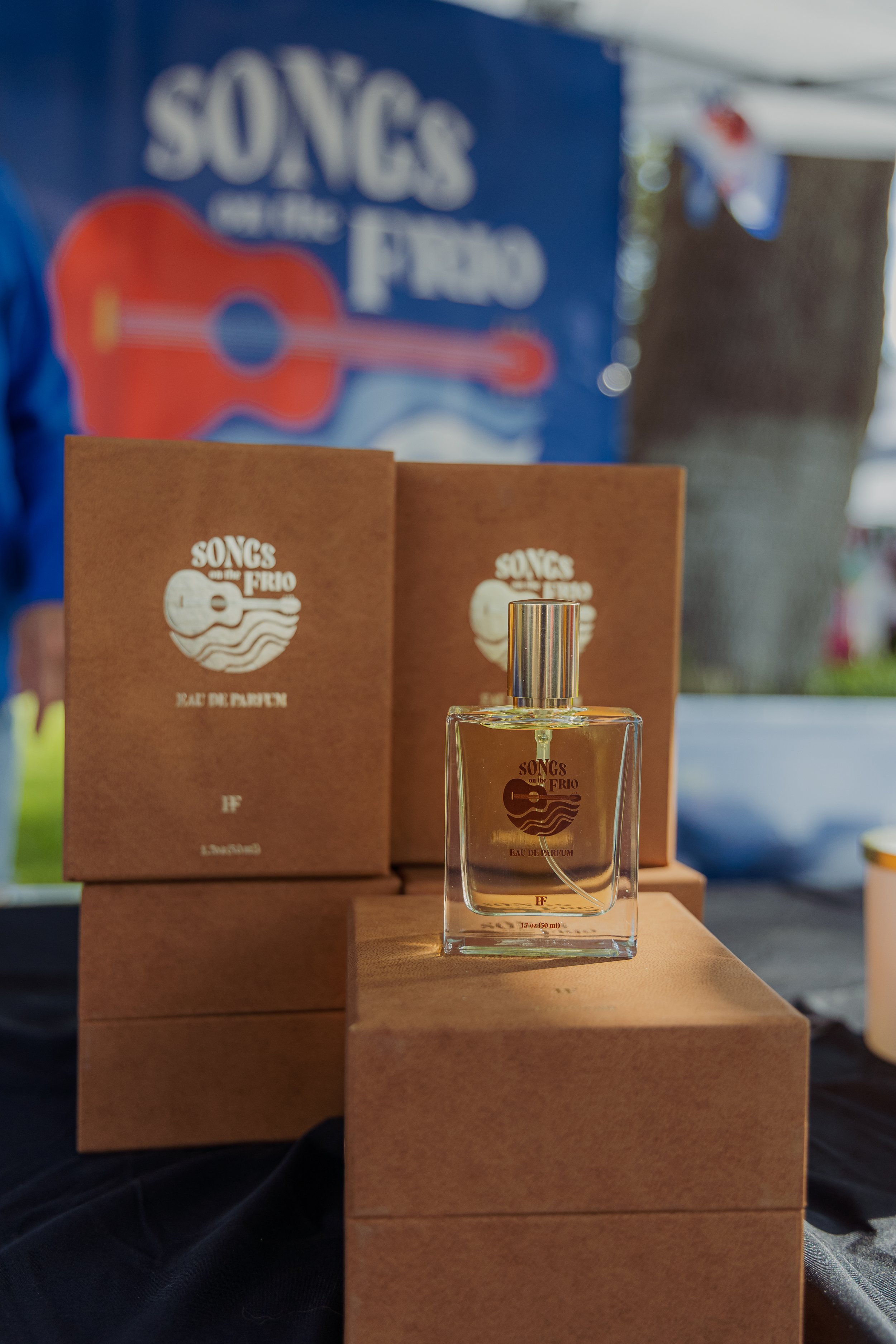 A bottle of SONGS OF THE FRIO perfume displayed on a wooden block with its packaging behind it, set outdoors with a blurred background featuring a blue sign with a guitar and the text 'SONGS OF THE FRIO'.