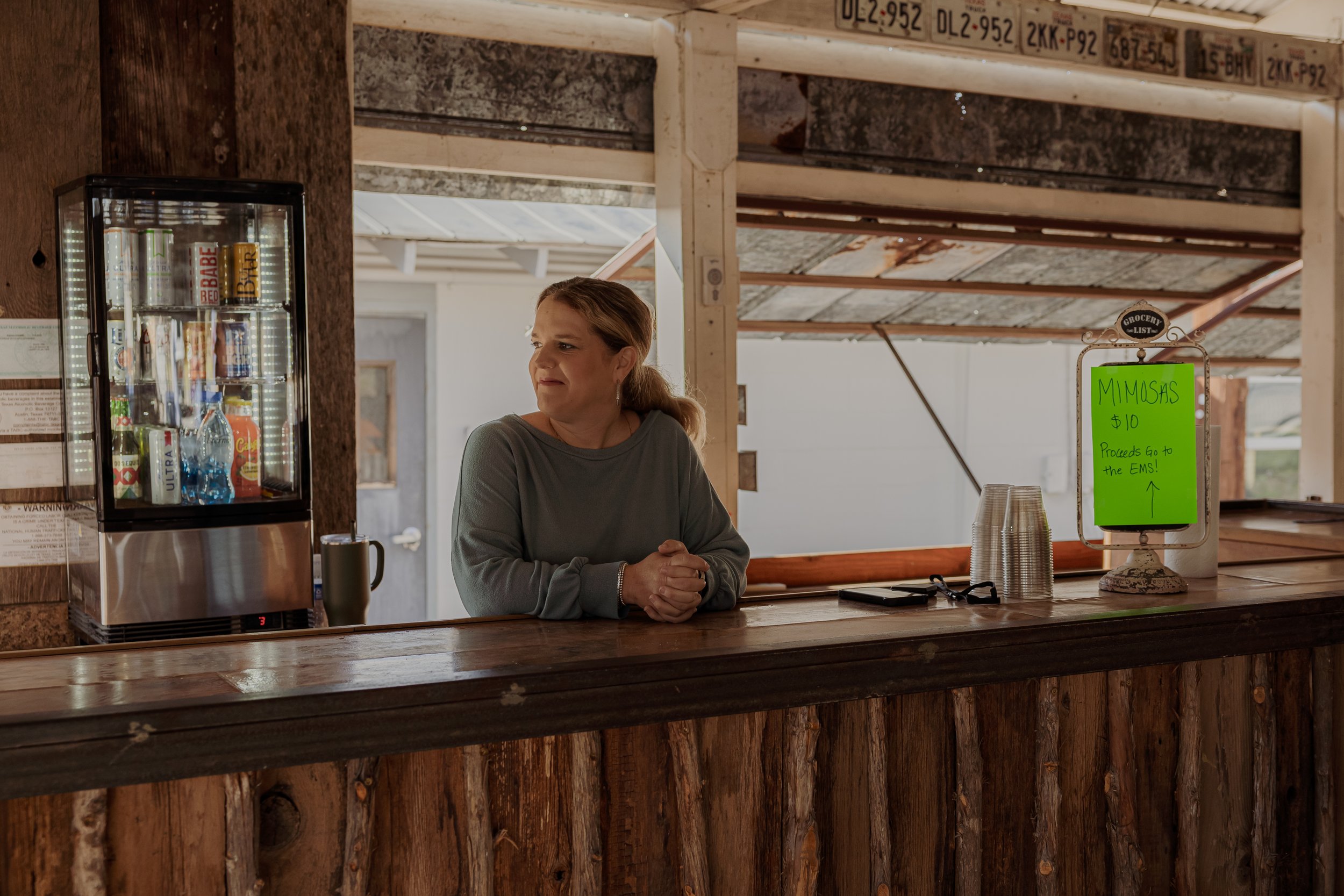 A woman standing behind a rustic wooden counter at a small stand with a bright green sign promoting mimosas for ten dollars, with proceeds going to the EMS. There is a refrigerator with bottled drinks to her left and a collection of disposable cups a