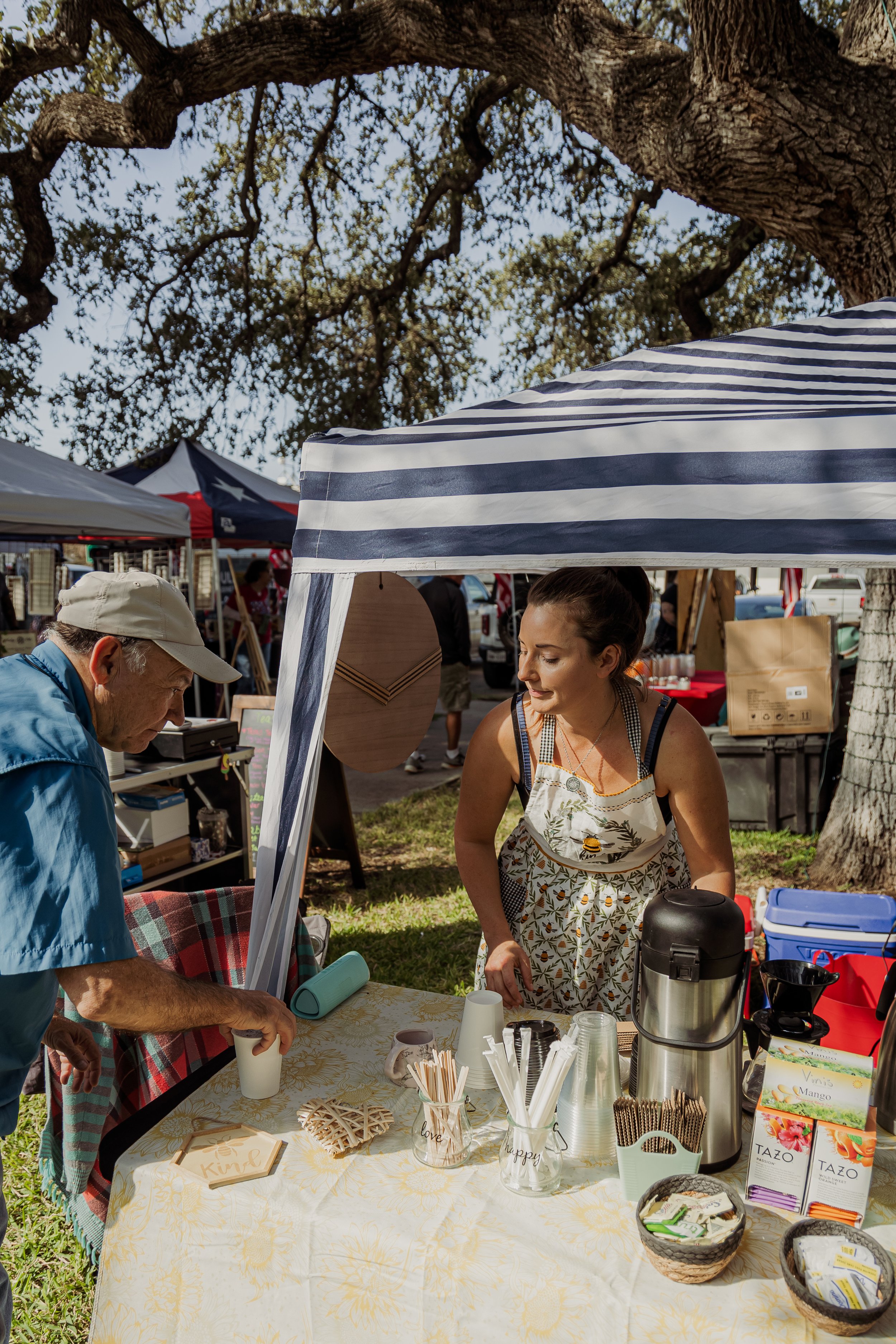 A woman serving a drink to an older man at an outdoor market stall, with a striped canopy and trees in the background.