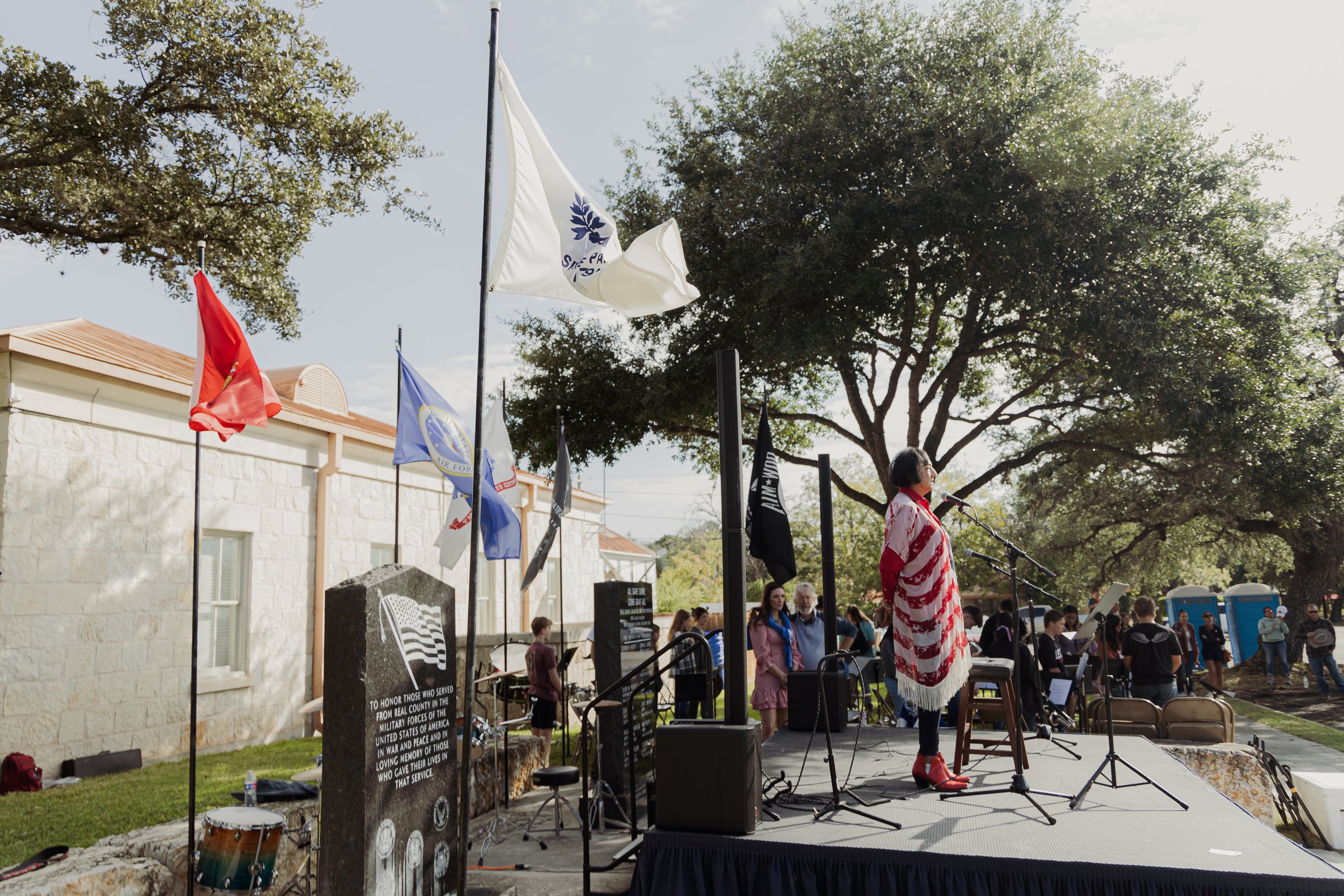 A woman dressed in a red and white American flag shawl speaks into a microphone on an outdoor stage, with people gathered around. Several flags, including American and military flags, are displayed on poles. There is a memorial stone with an inscript
