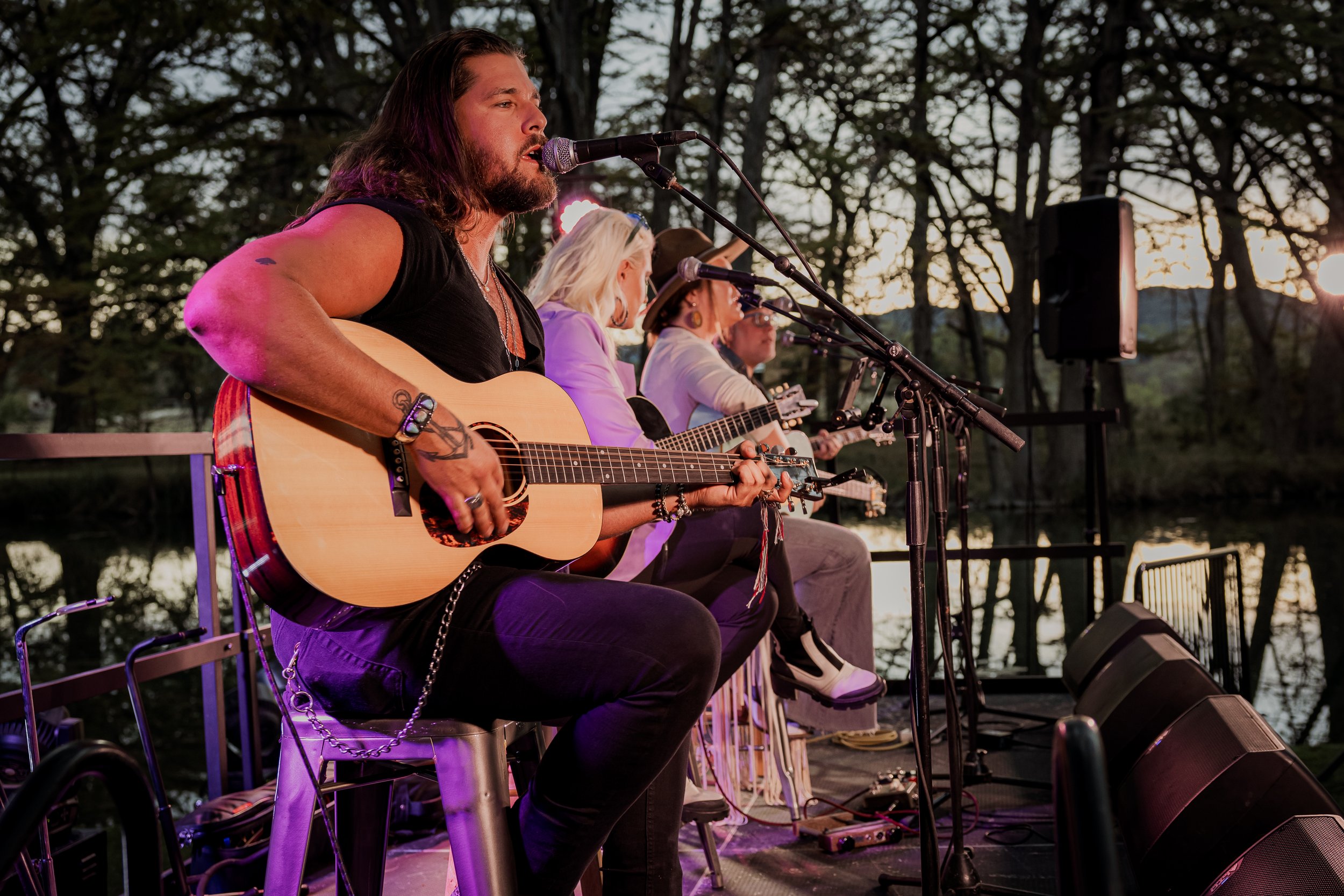 A group of musicians performing outdoors near a body of water at sunset, with three women and one man playing guitars and singing into microphones.