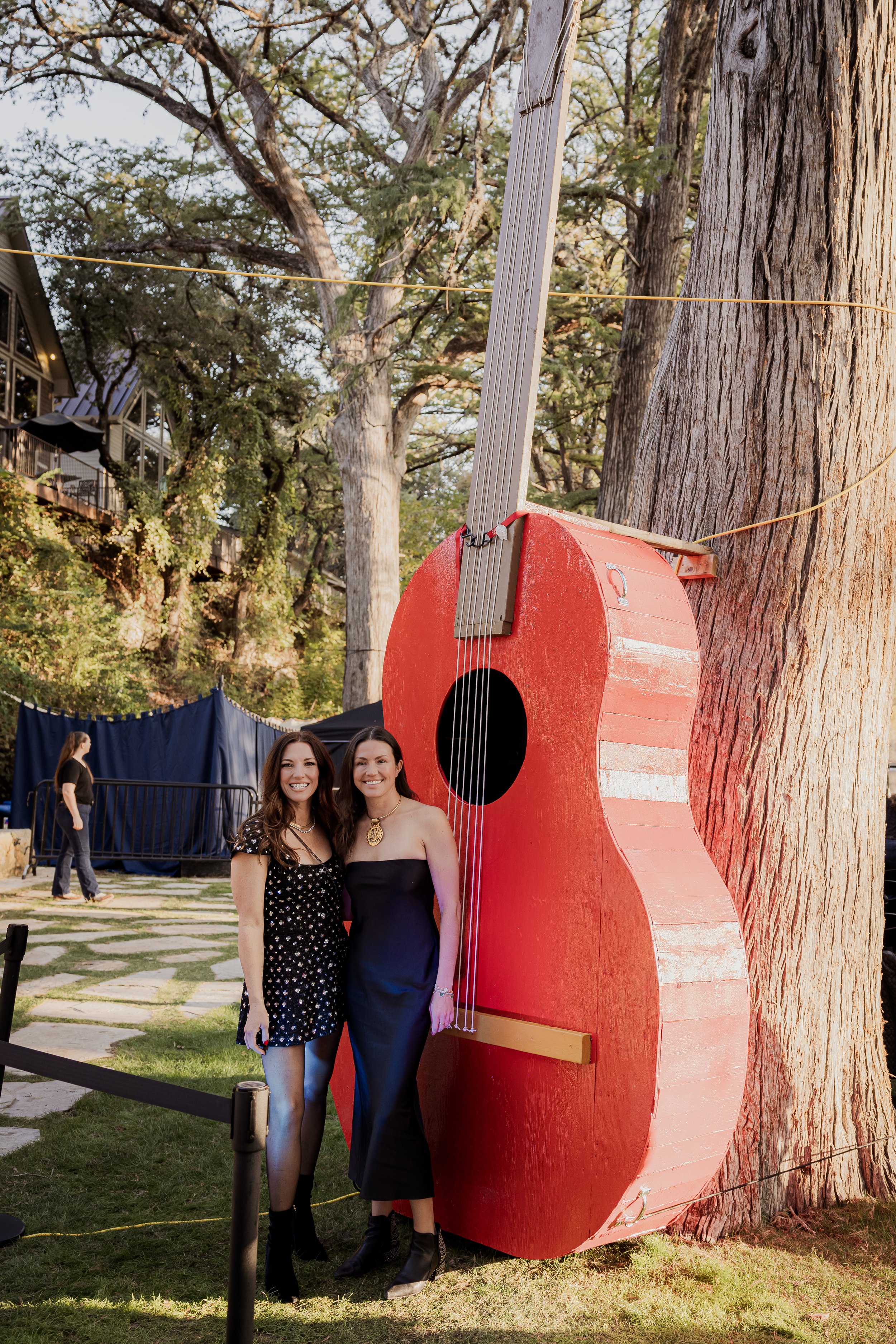 Two women standing outdoors beside a large red guitar-shaped sculpture, with trees and a building in the background.