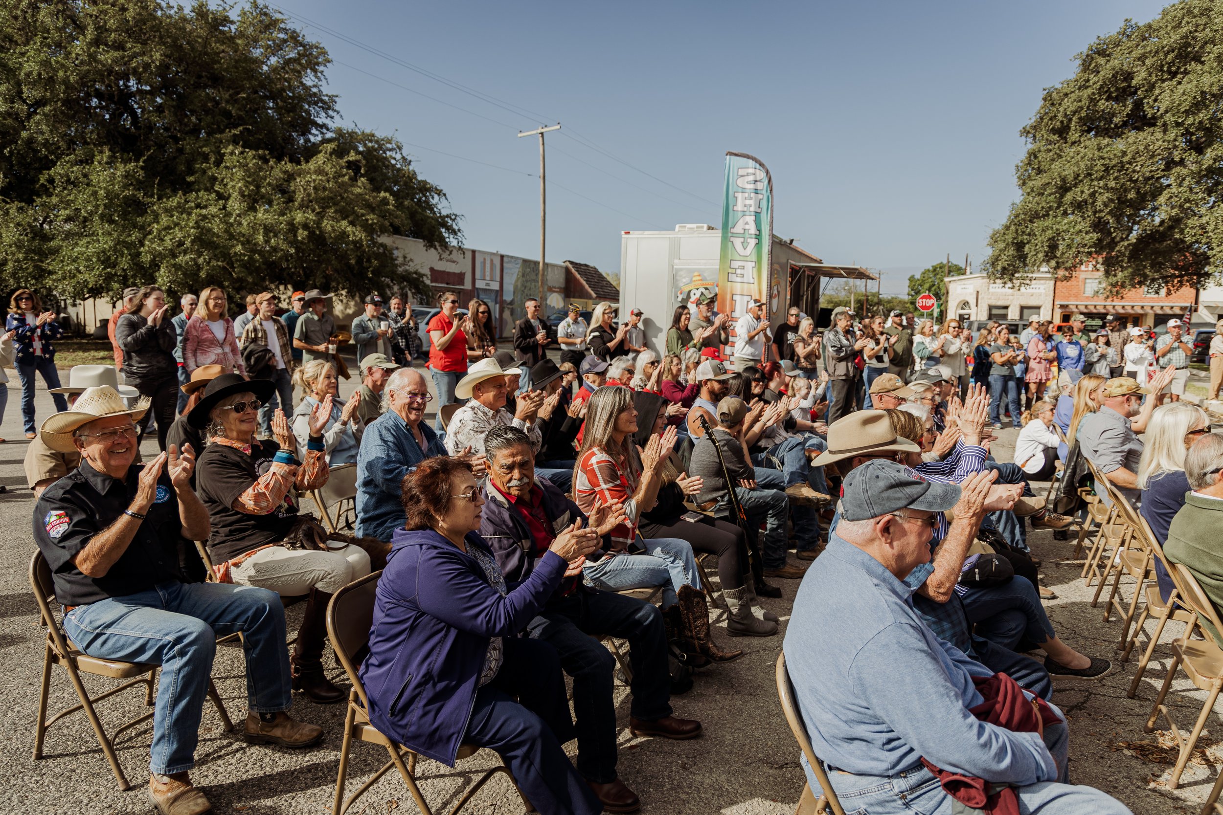 Crowd of people enjoying an outdoor event on a sunny day, sitting and standing on a street with trees and buildings in the background.