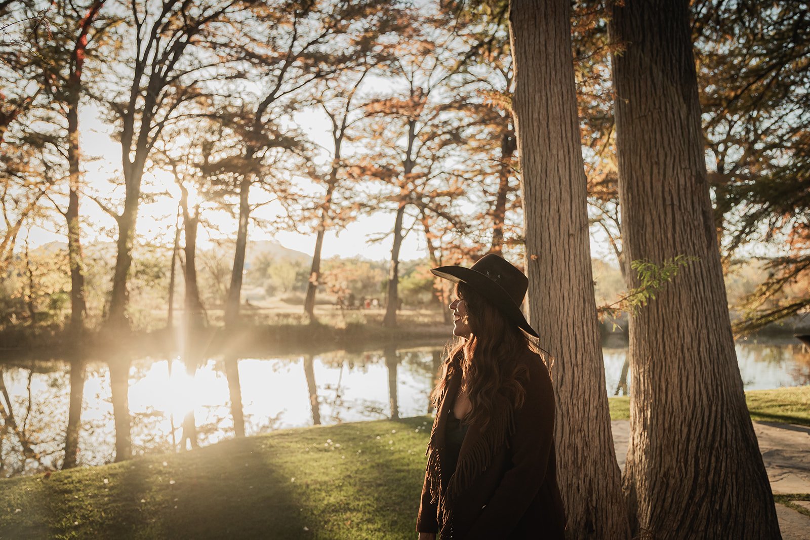 A woman with long wavy hair wearing a black hat and dark jacket stands beside a tree near a body of water during sunset, with trees and sunlight in the background.
