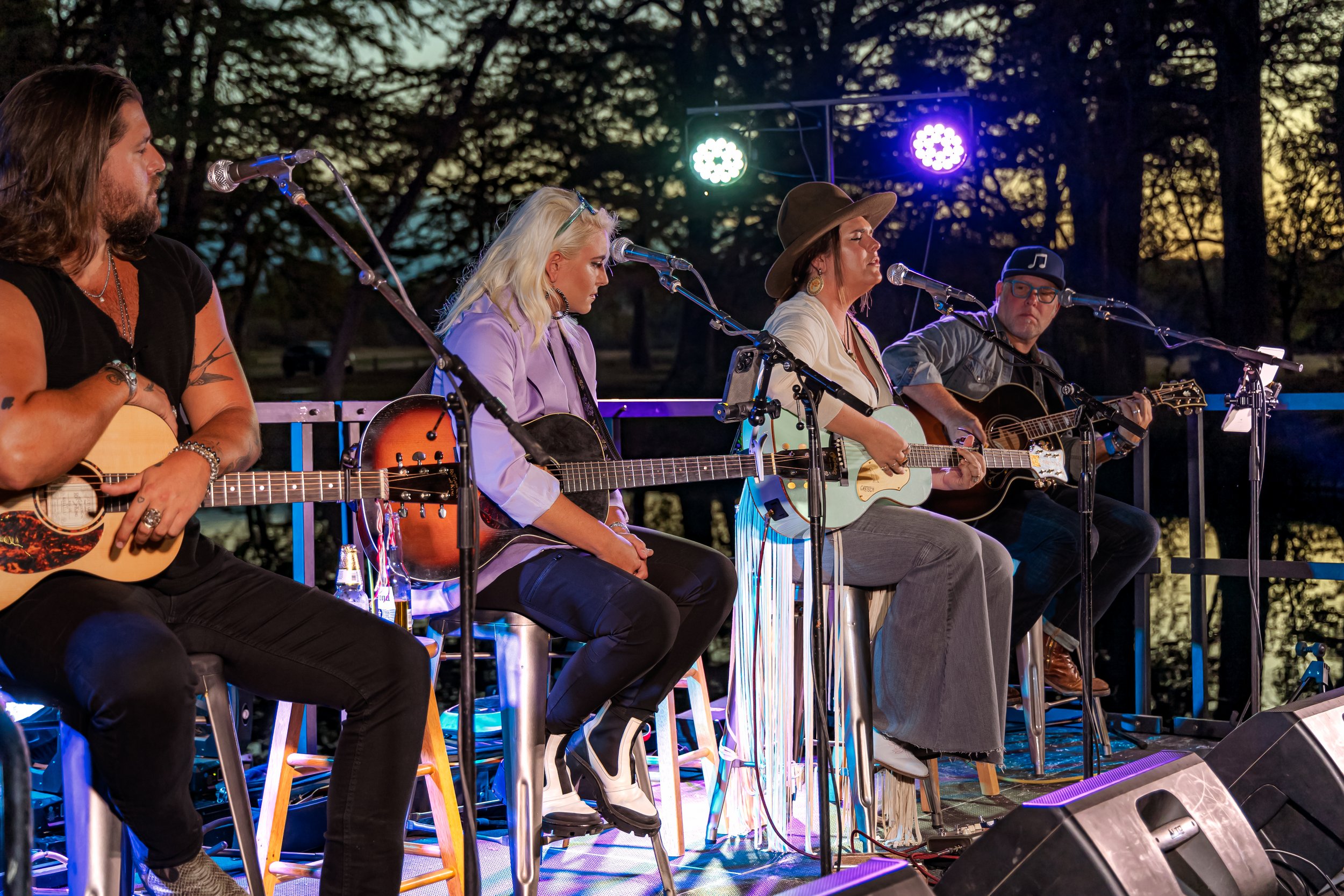 Five musicians playing acoustic guitars on an outdoor stage during sunset, with colorful stage lights behind them.