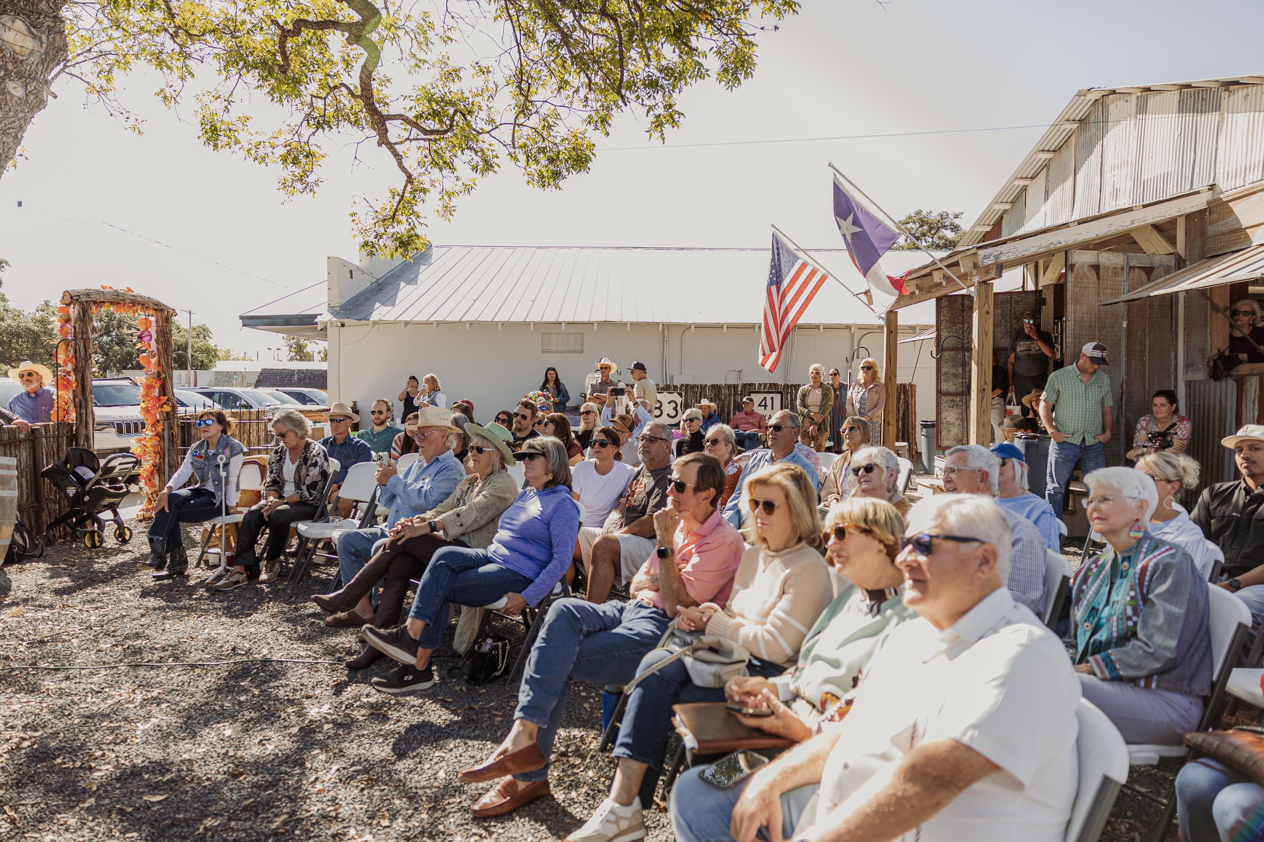 A group of people sitting outdoors on folding chairs, attending an event, with some wearing sunglasses and hats, under a tree, in front of a rustic building with flags and parked cars nearby.