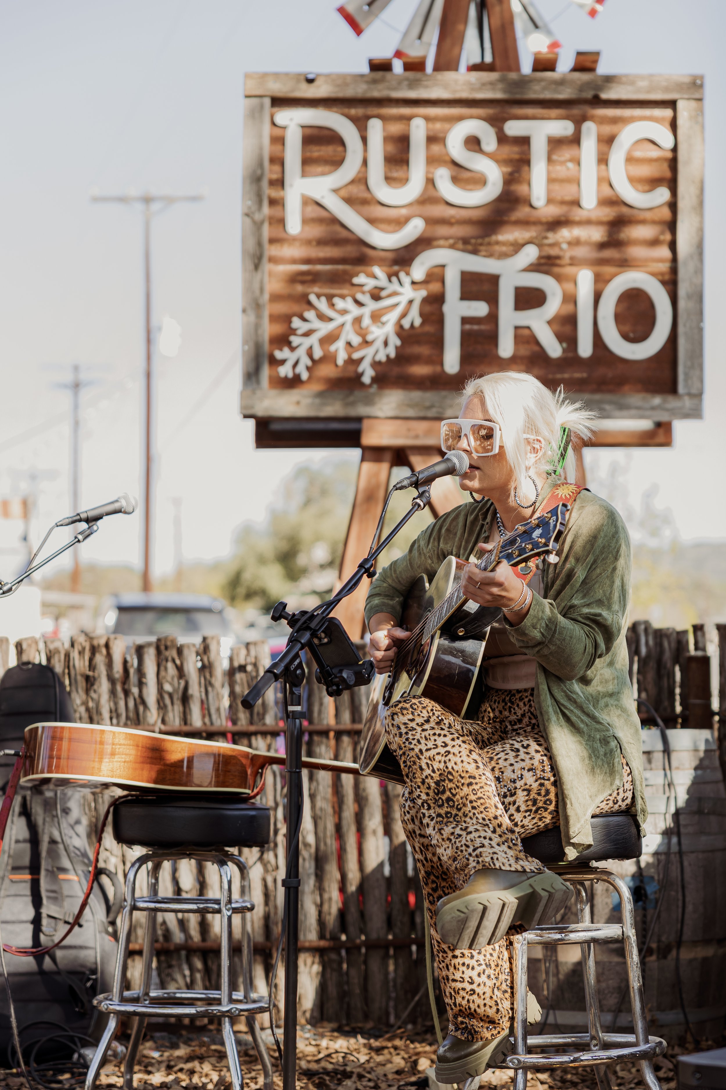 A woman with blonde hair, wearing large sunglasses, a green jacket, and leopard print pants, plays an acoustic guitar while singing into a microphone at an outdoor performance with a rustic sign that says "Rustic Rio" behind her.