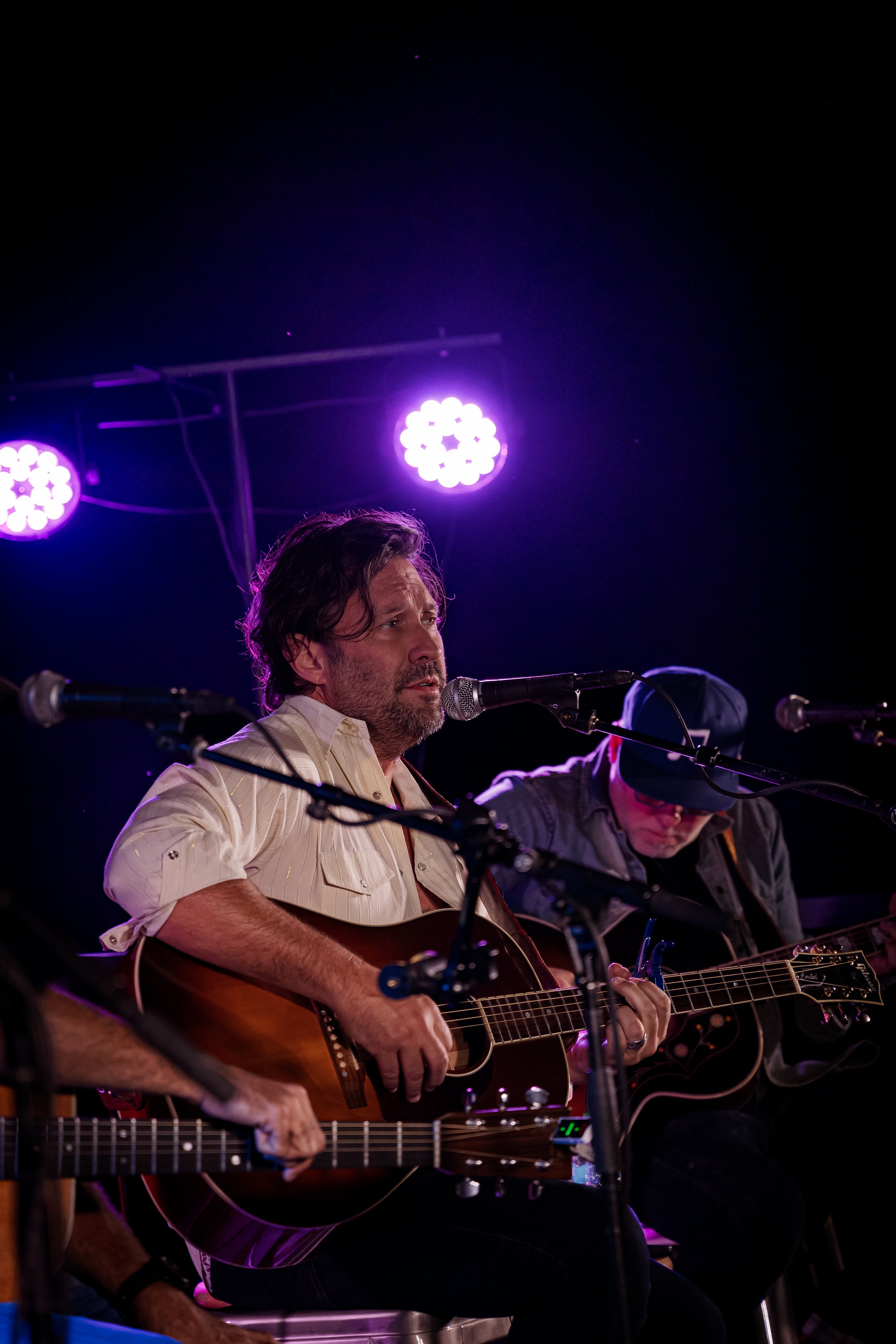 Musicians performing live on stage at night under purple stage lights, playing acoustic guitars and singing into microphones.