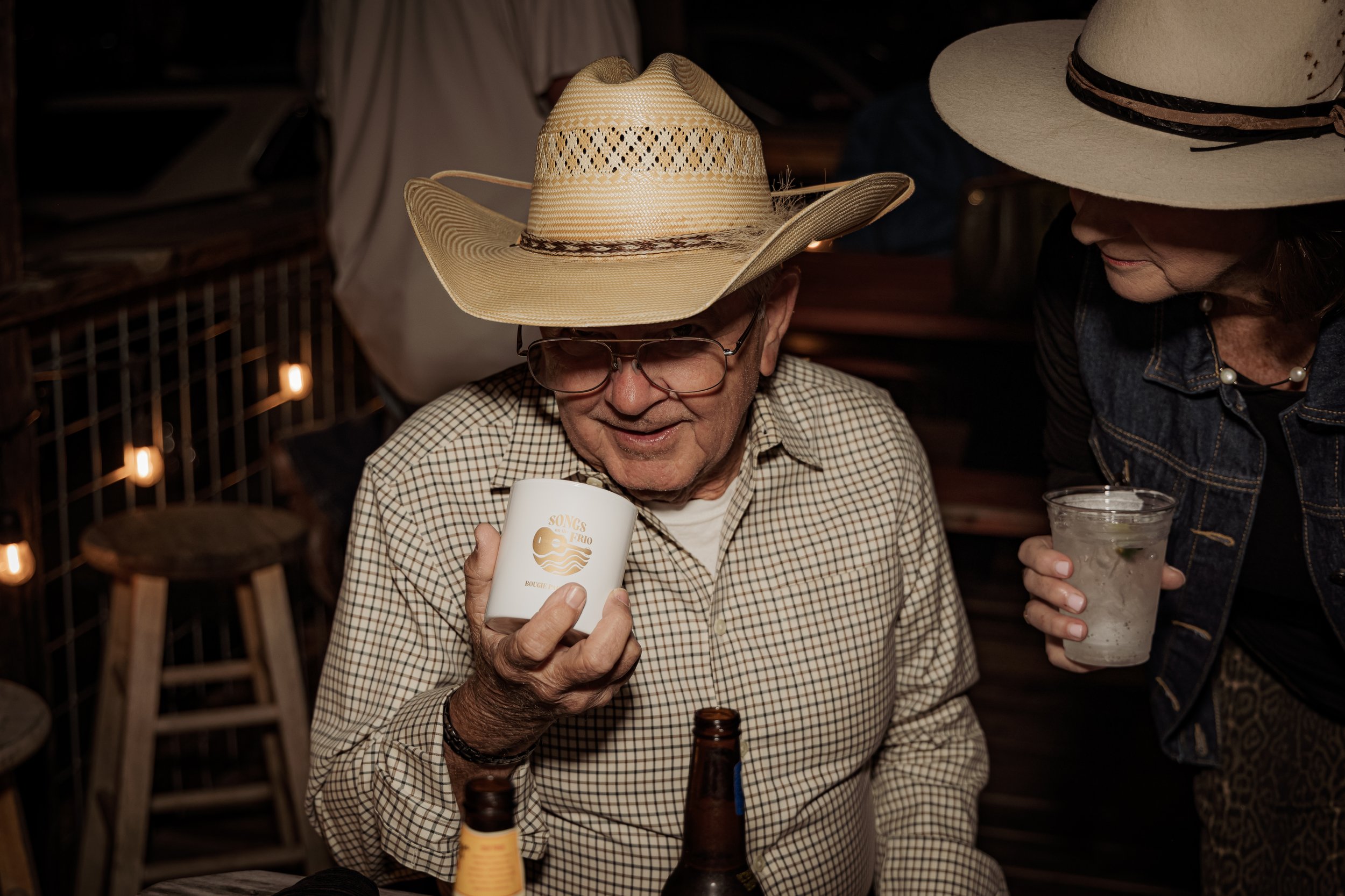 An elderly man wearing a cowboy hat, glasses, and a checkered shirt holds a white mug, while a woman next to him in a wide-brimmed hat and denim jacket holds a clear glass with a drink. They are sitting at a wooden bar with stools, in a cozy, dimly-l