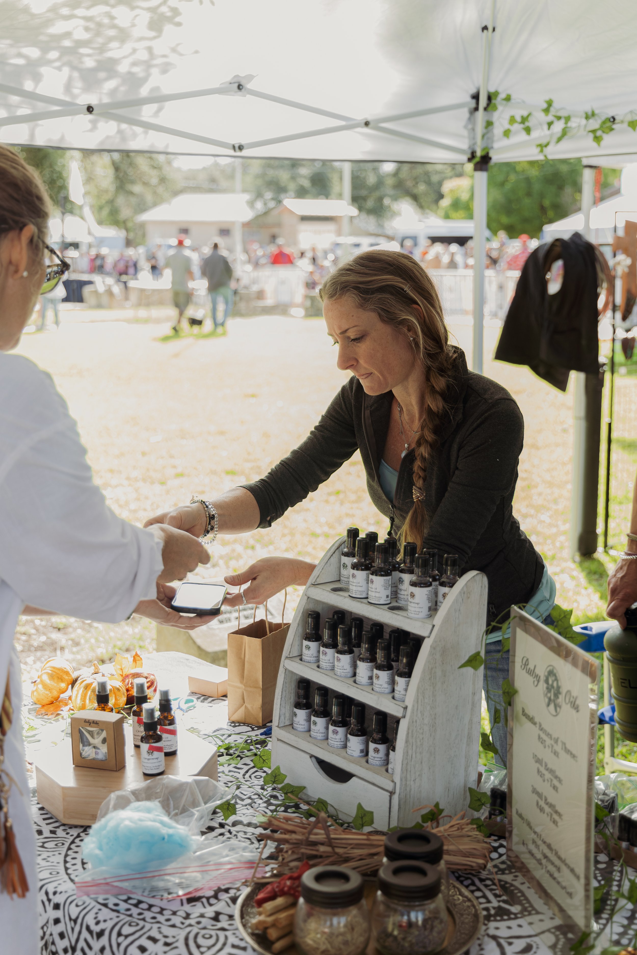 A woman selling essential oils at an outdoor market stall during the day, with various bottles on display and a customer making a purchase.