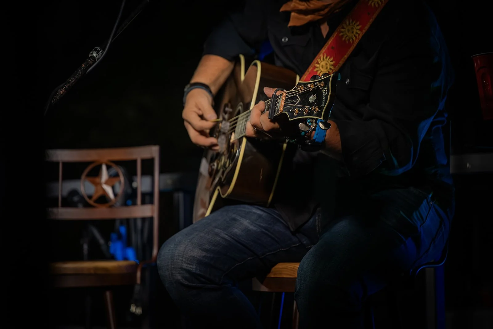 A person playing an acoustic guitar on stage, with a chair and musical equipment nearby, in a dark setting.