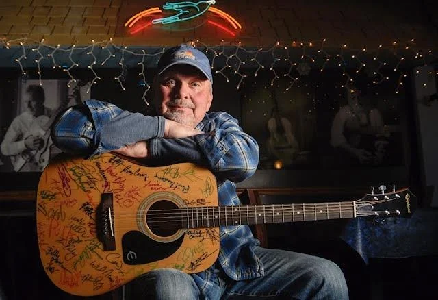 A man with a baseball cap sitting with arms resting on an acoustic guitar covered in signatures and messages in a dimly lit bar or music venue.