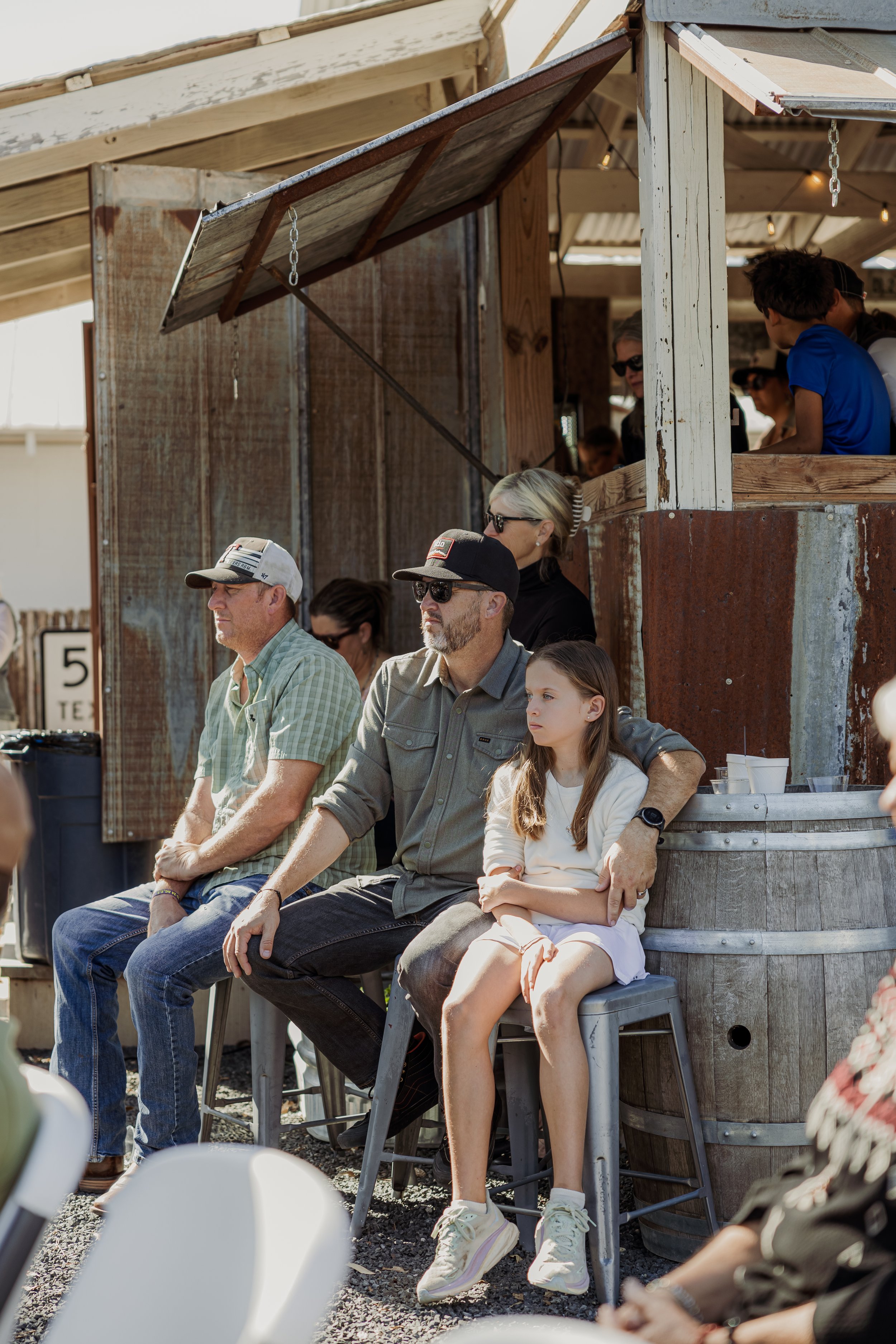 People sitting and watching at an outdoor event, some wearing hats and sunglasses, with a rustic wooden and metal structure in the background.