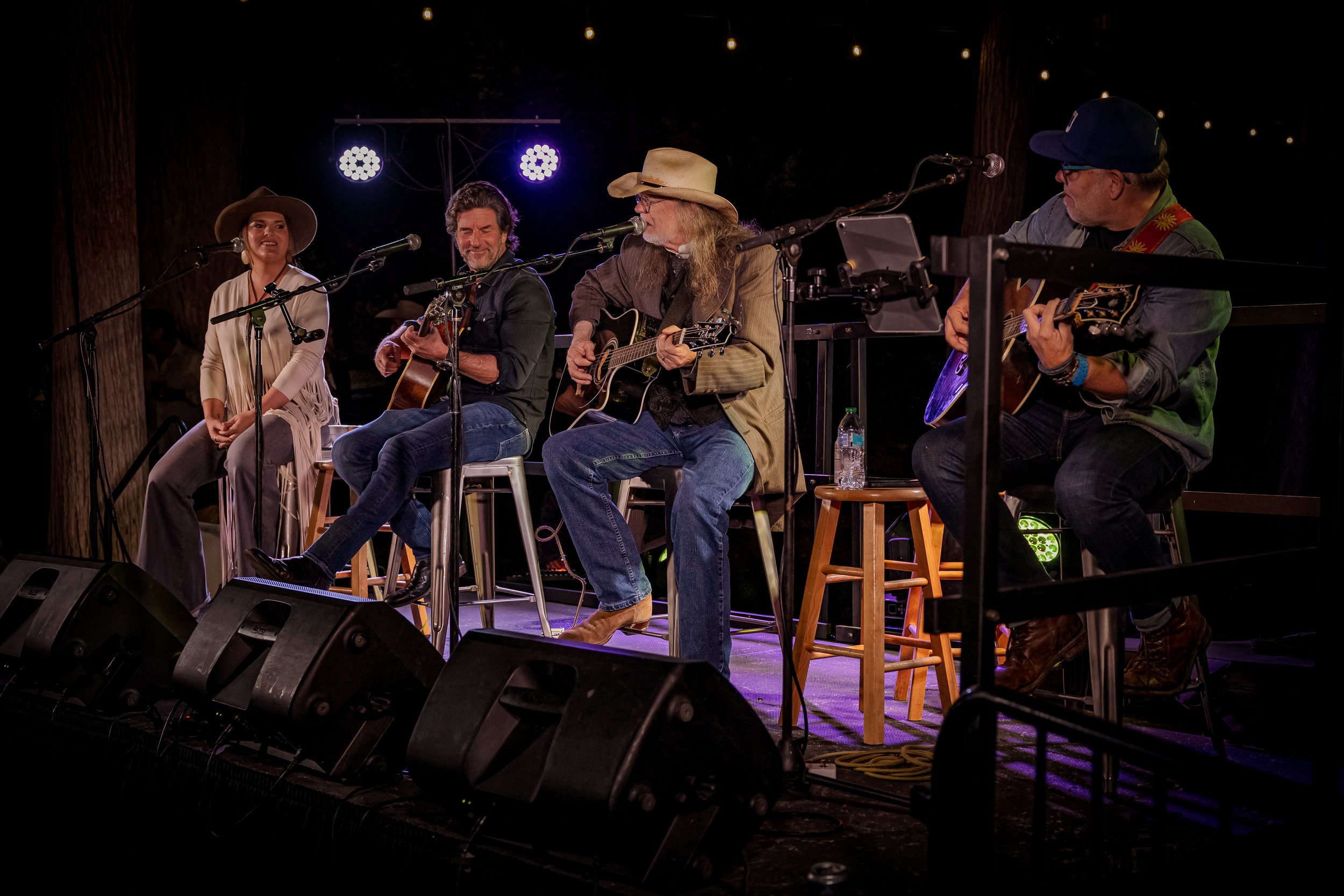 Four musicians performing on stage with guitars and vocals, under string lights, with music equipment and monitors in front.