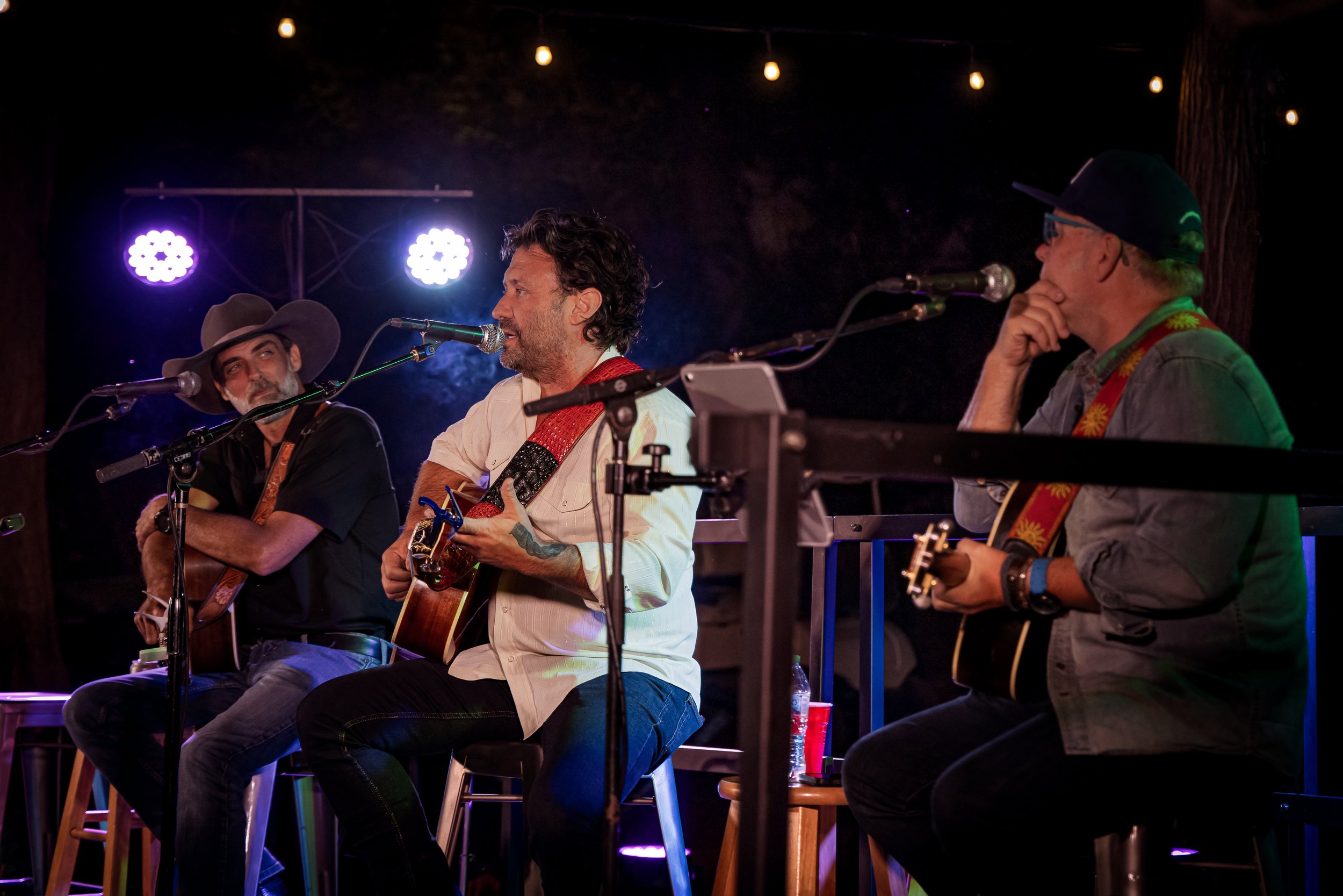 Three musicians sitting on stools playing acoustic guitars on stage under string lights at night, with a dark background and two bright stage lights shining from behind.