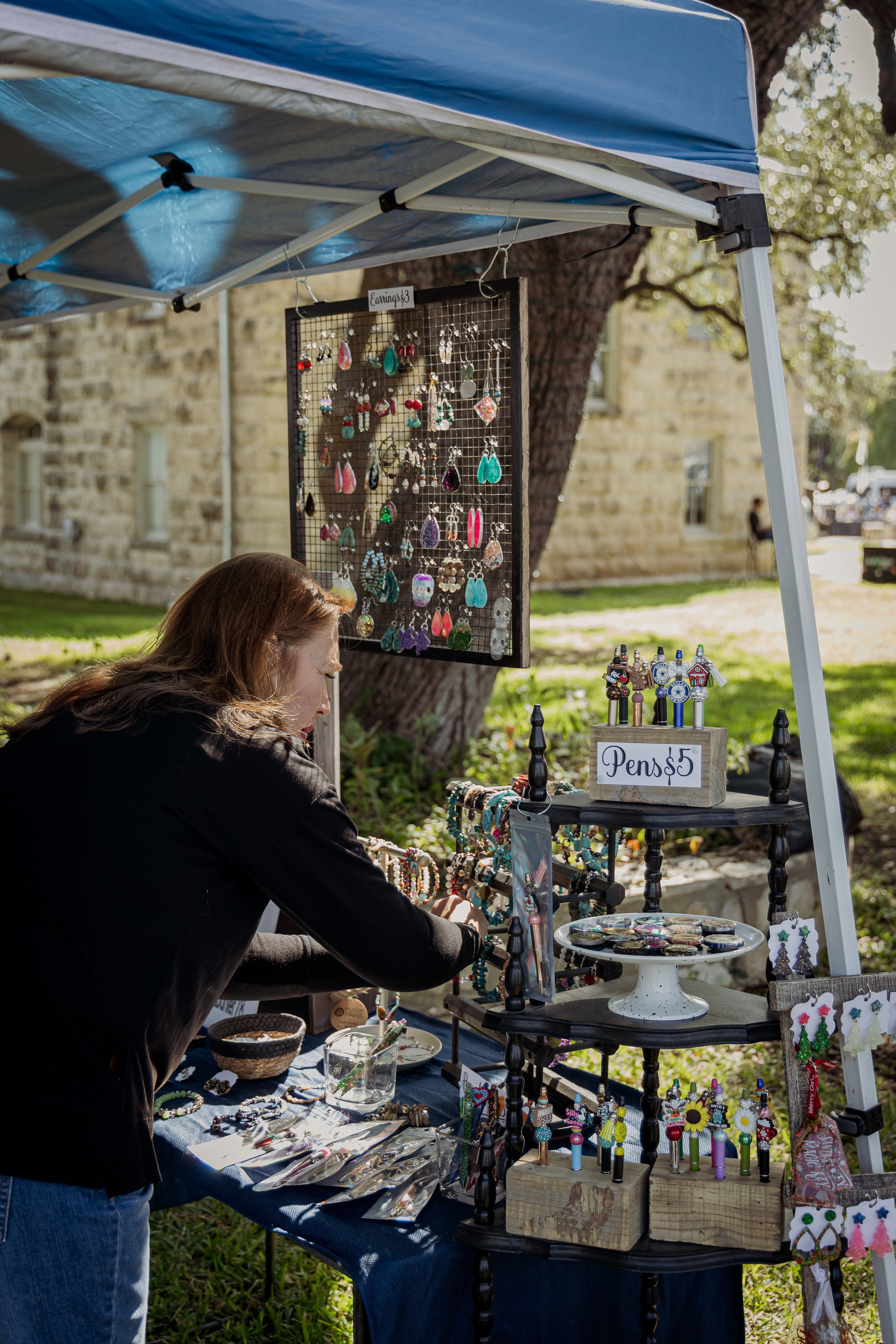 A woman are browsing handcrafted jewelry at an outdoor market booth with a sign indicating earrings priced at $3 and pens priced at $5.