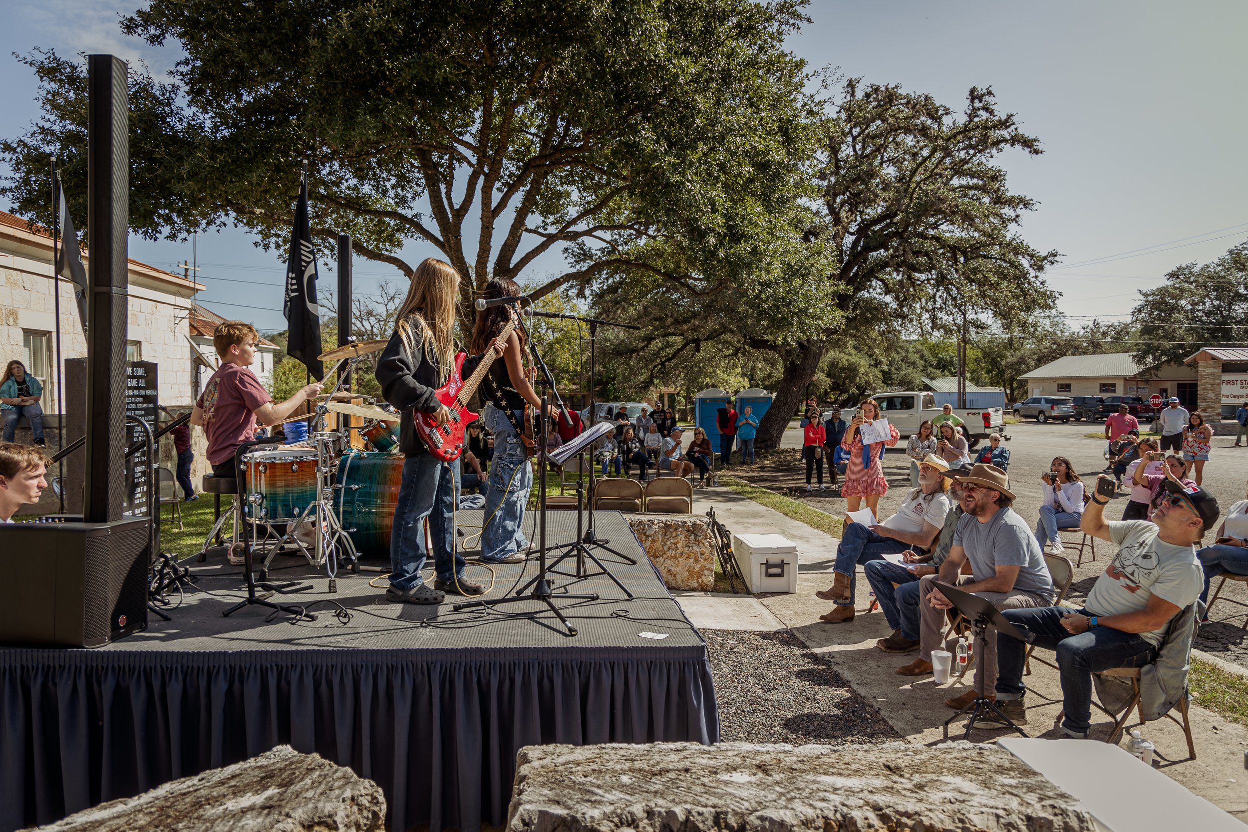A band of young musicians performs outdoors on a small stage with guitar, drums, and microphone, in front of an audience seated and standing under a large tree on a sunny day. People are using phones and taking photos, with some sitting on chairs and