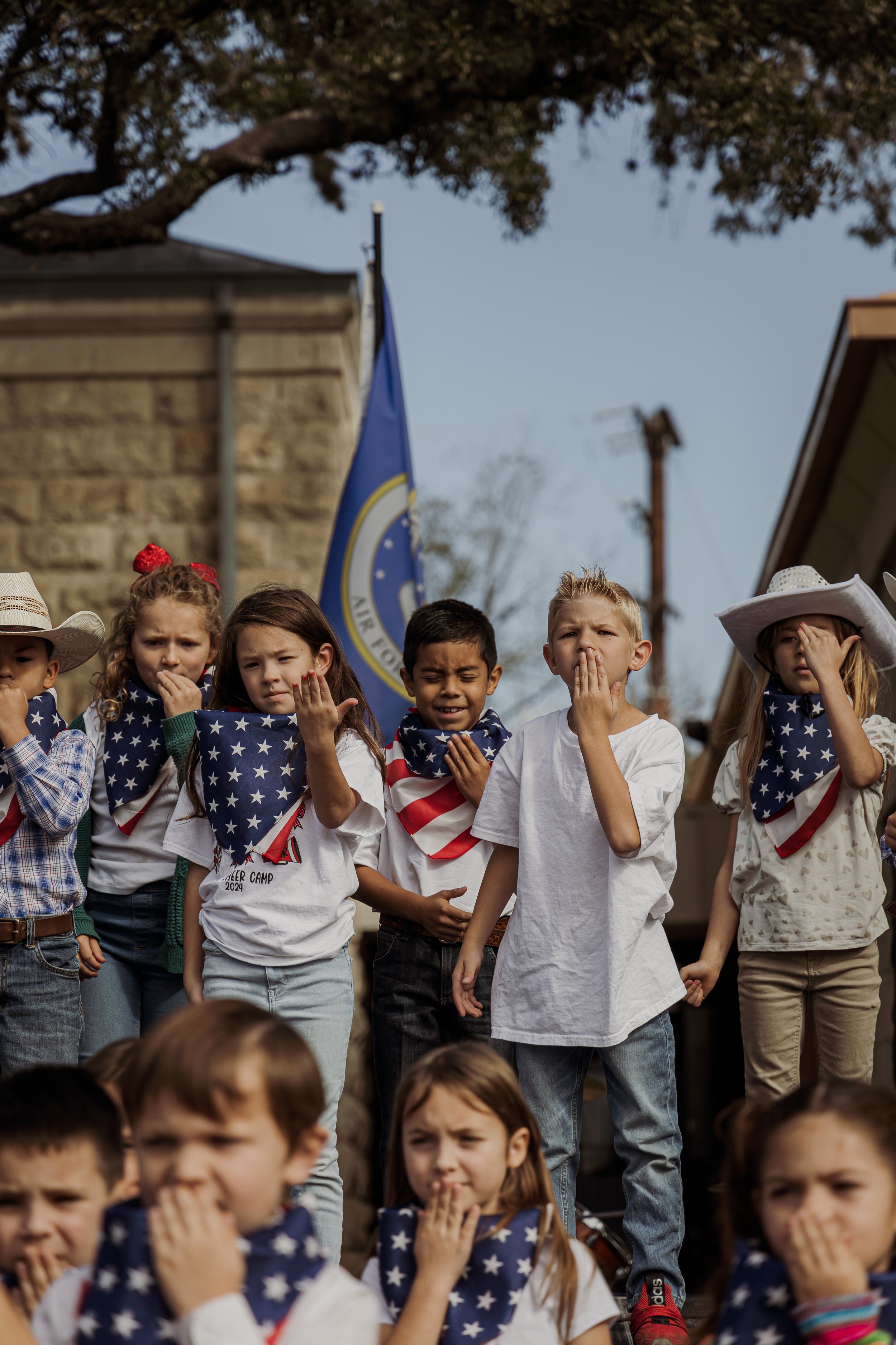 A group of children dressed in patriotic American-themed clothing and bandanas, holding their hands over their hearts, participating in a patriotic pledge or ceremony outdoors.