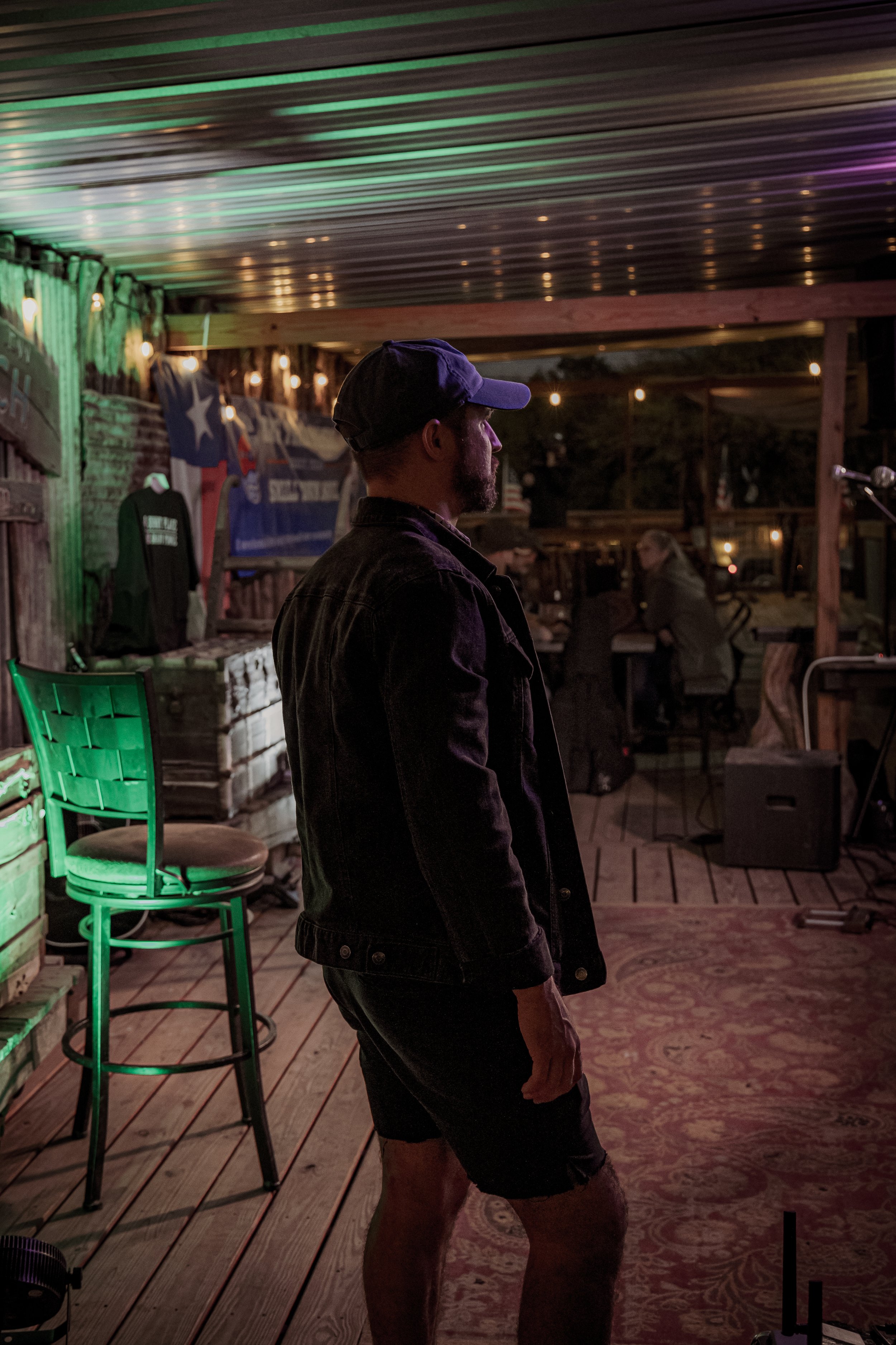 A man with a beard and cap standing in a barn-like setting with wooden flooring, decorative lighting, and people sitting at tables in the background.