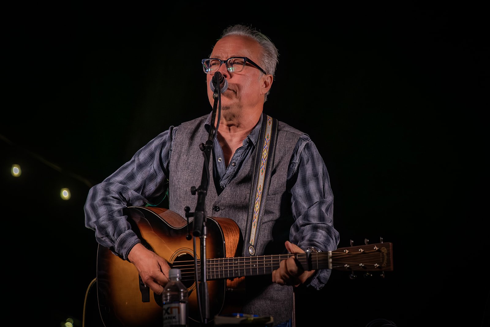 Man with glasses playing an acoustic guitar and singing into a microphone on a dark stage.
