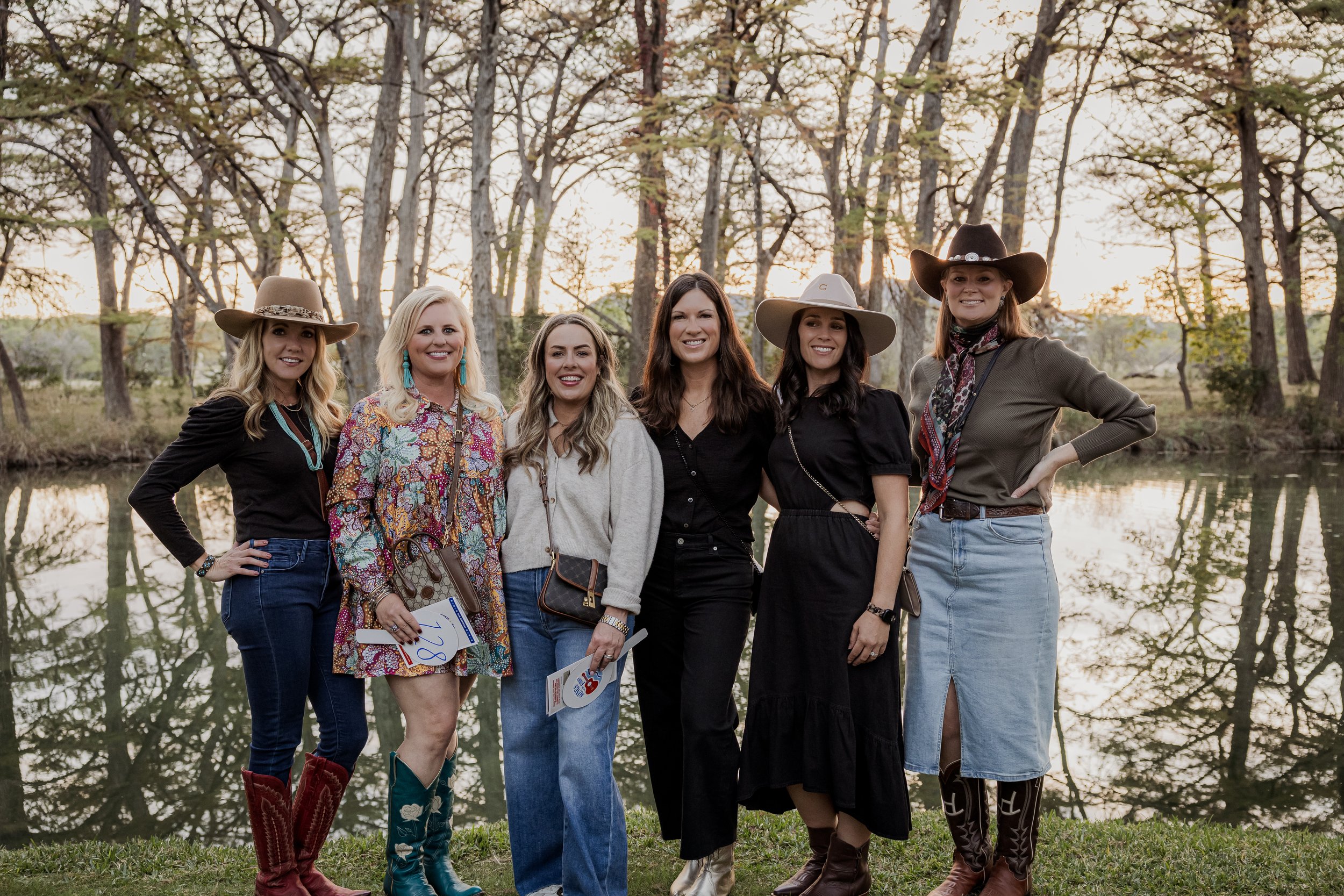 Six women standing outdoors near the Frio River, dressed in country or western style, with trees in the background at sunset.