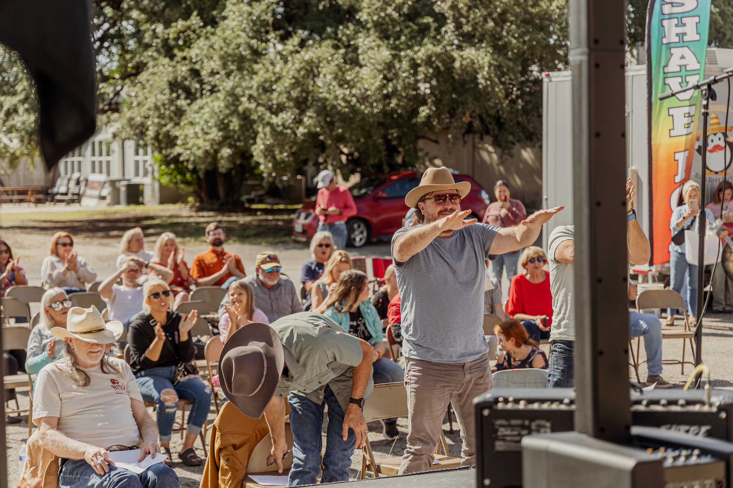 A man wearing a cowboy hat and sunglasses stands with arms outstretched in front of an outdoor audience at a sunny event, with several people seated and clapping behind him.