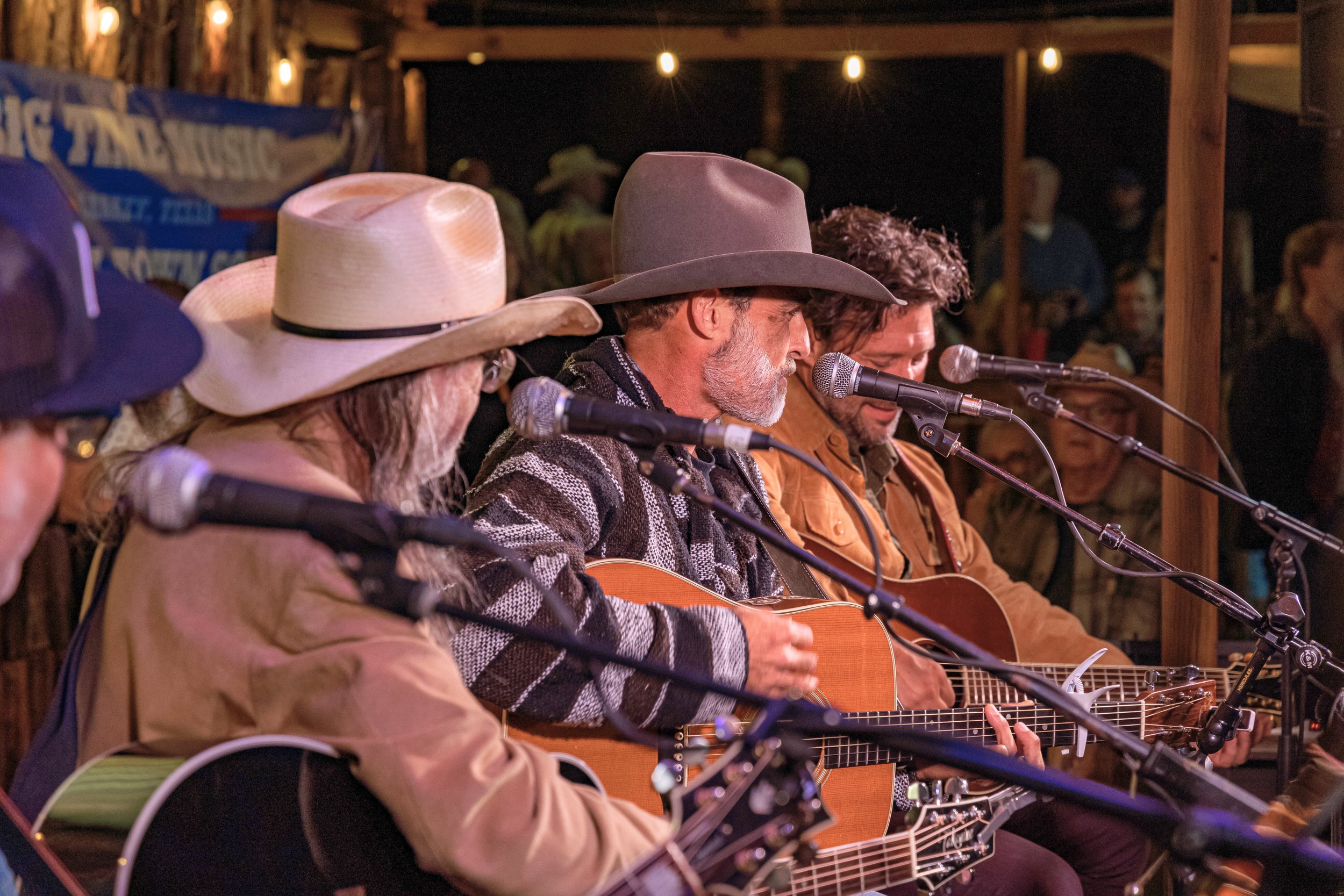A group of musicians playing guitars and singing into microphones at an outdoor concert during nighttime, with string lights overhead and people in the background.