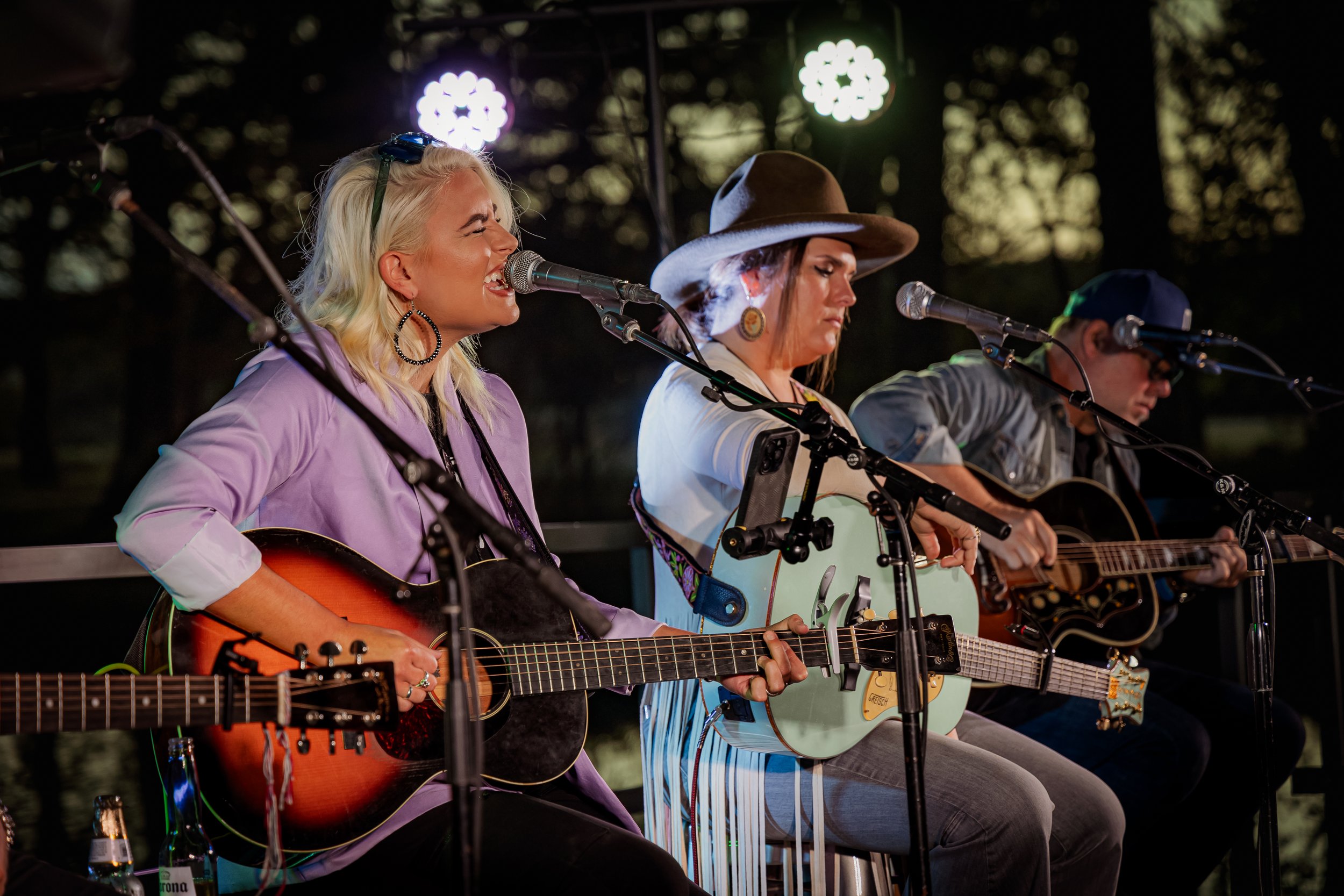 Three musicians sitting on stools performing with guitars on a stage with microphones, at an outdoor event during evening time.