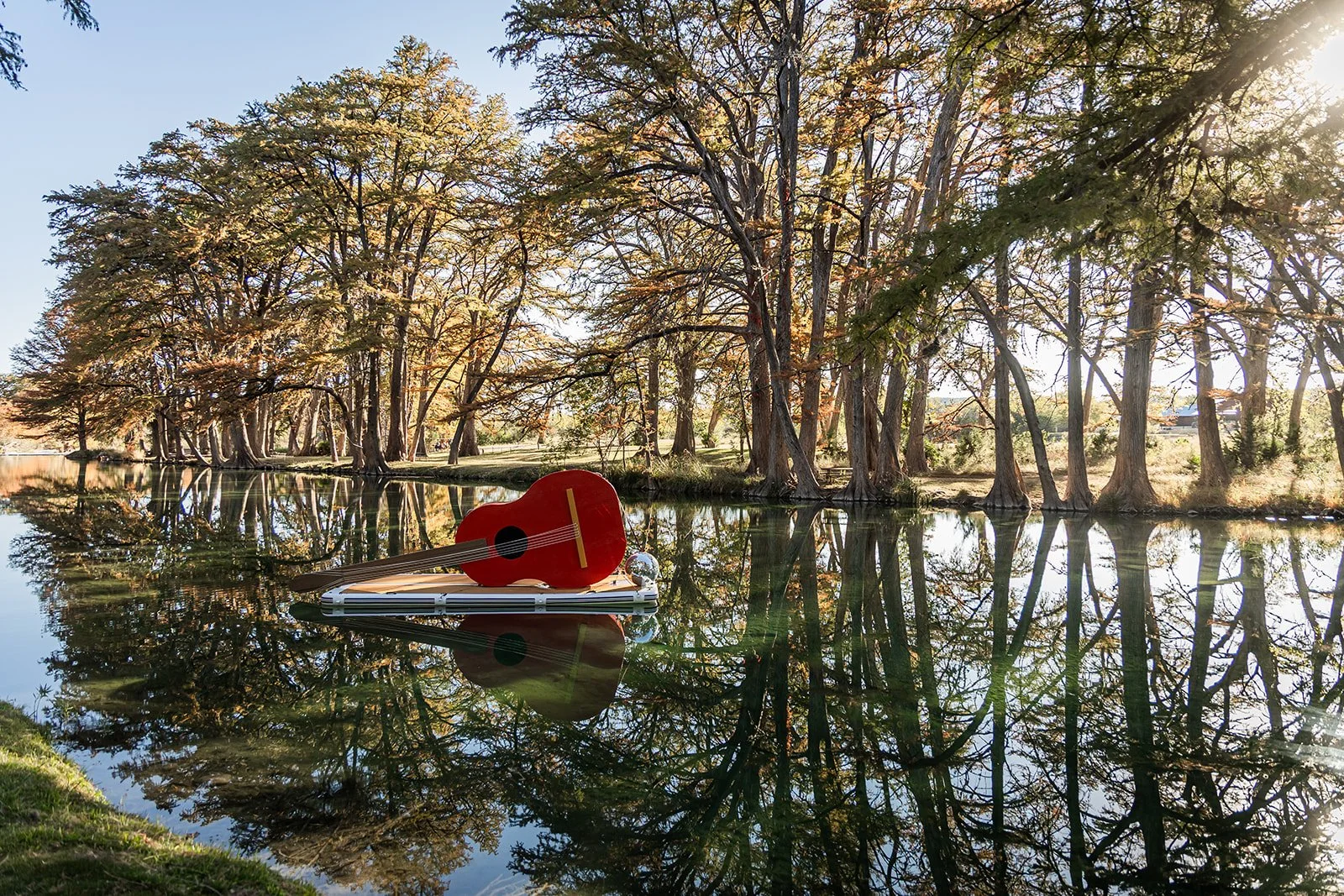 A small red guitar and a folded white stand-up paddleboard lying on calm water, with tall trees and their reflections visible in the water, and sunlight shining through the trees.