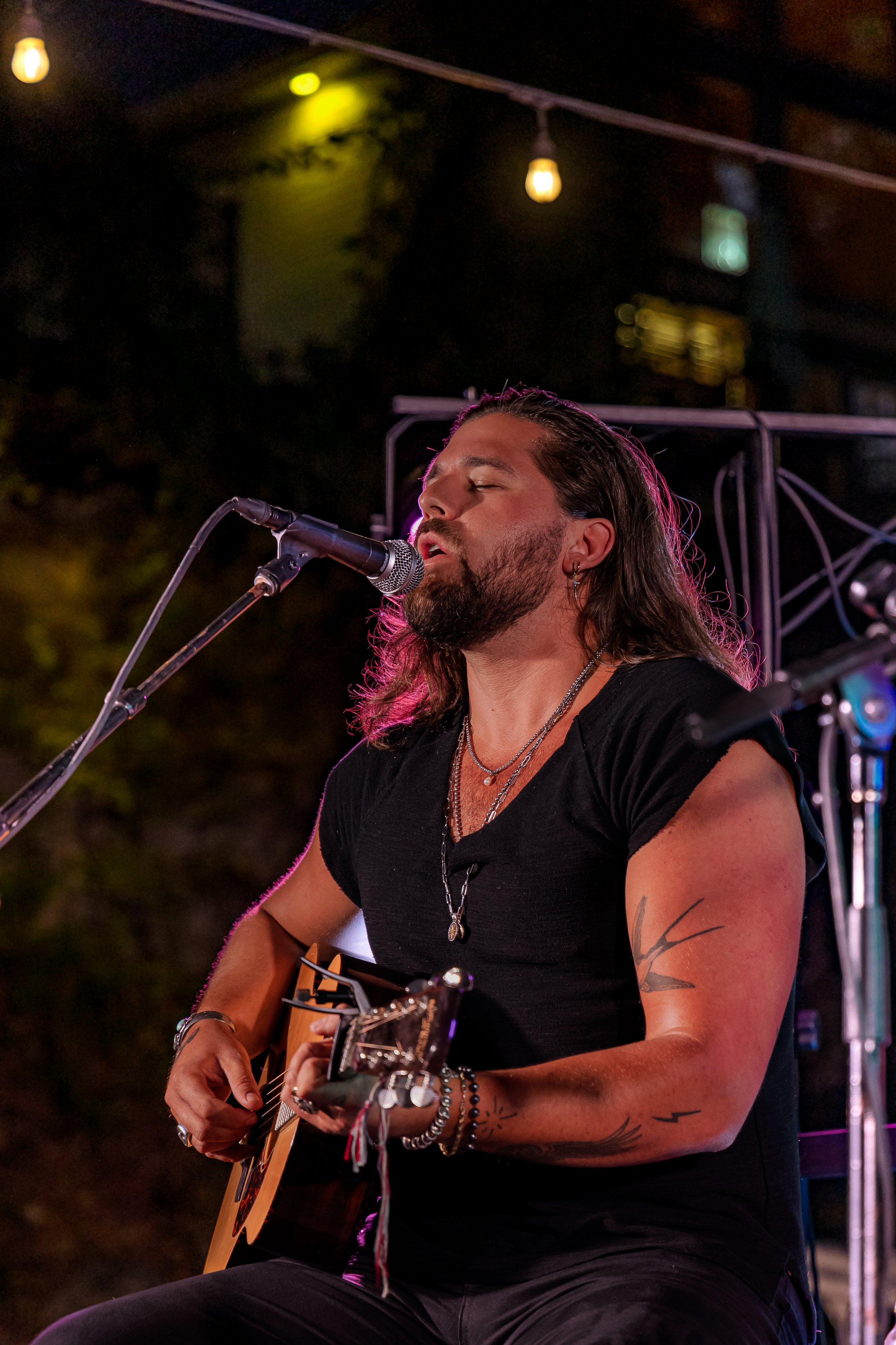 A male musician with long hair and tattoos, wearing a black t-shirt, plays an acoustic guitar and sings into a microphone during a live performance at night, illuminated by stage lights and hanging bulbs.