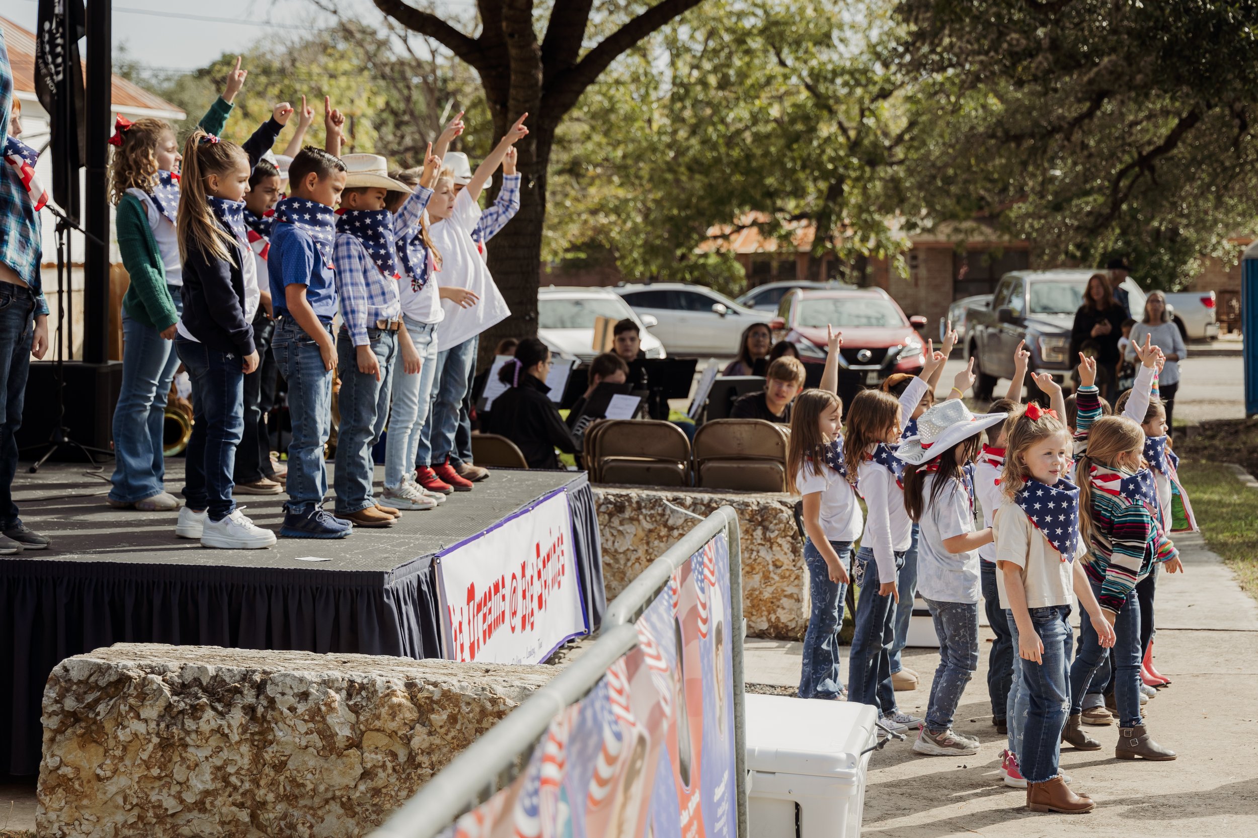 Children performing on an outdoor stage and in front of the stage during a patriotic event, wearing outfits and bandanas with American flag patterns.