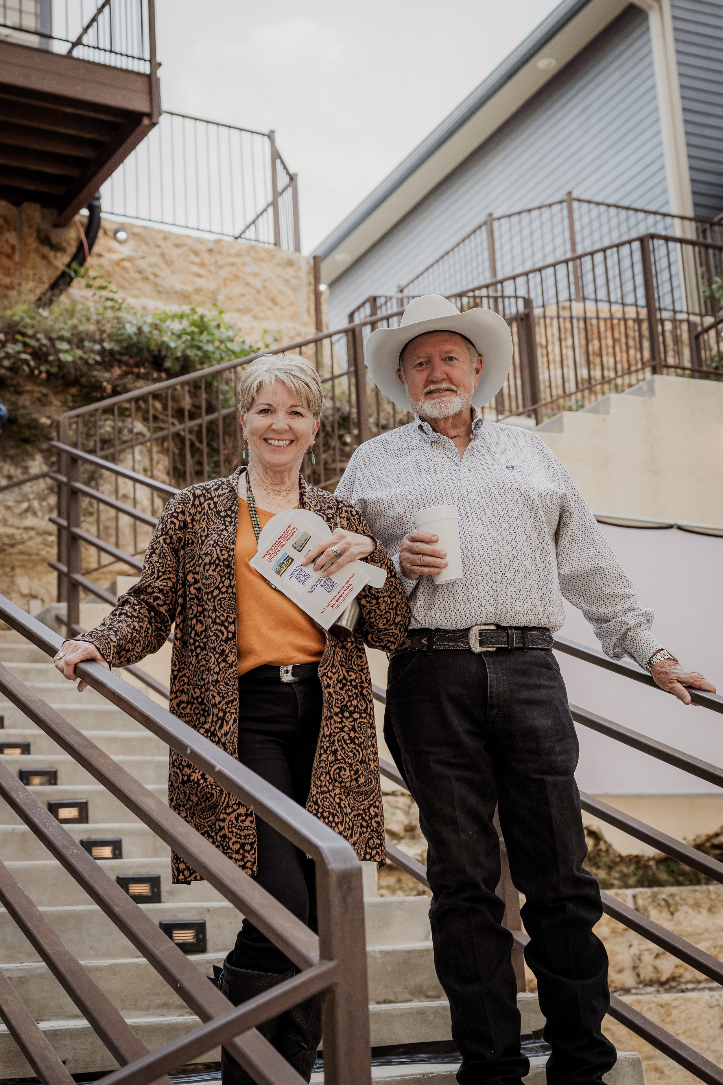 Older woman and man standing on outdoor staircase, smiling, with the woman holding a brochure and the man holding a cup, both dressed in casual attire, the woman wearing a patterned animal print coat and the man wearing a cowboy hat.