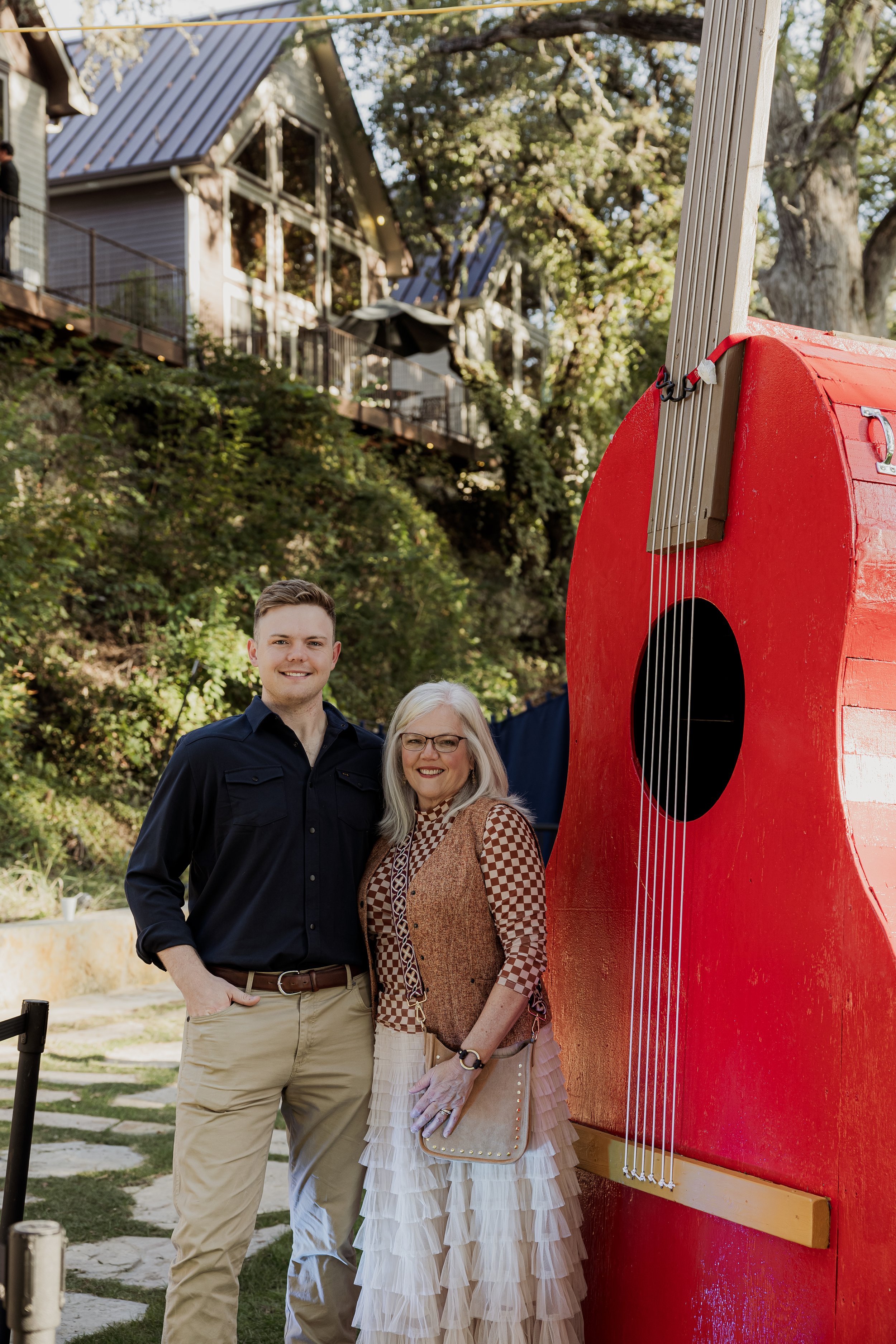 A young man and an older woman standing outdoors next to a large red guitar-shaped sculpture, smiling at the camera with trees and a house in the background.