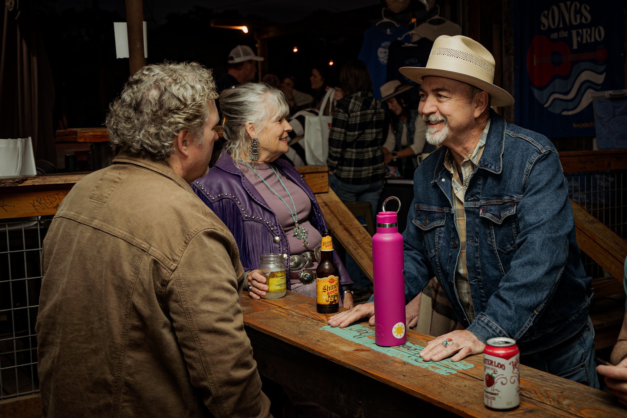 Three older adults, two men and a woman, talking and laughing at a wooden bar. The man on the right wears a straw hat and denim jacket, and the woman has long gray hair and wears a purple jacket. The man on the left has curly gray hair and a tan jack