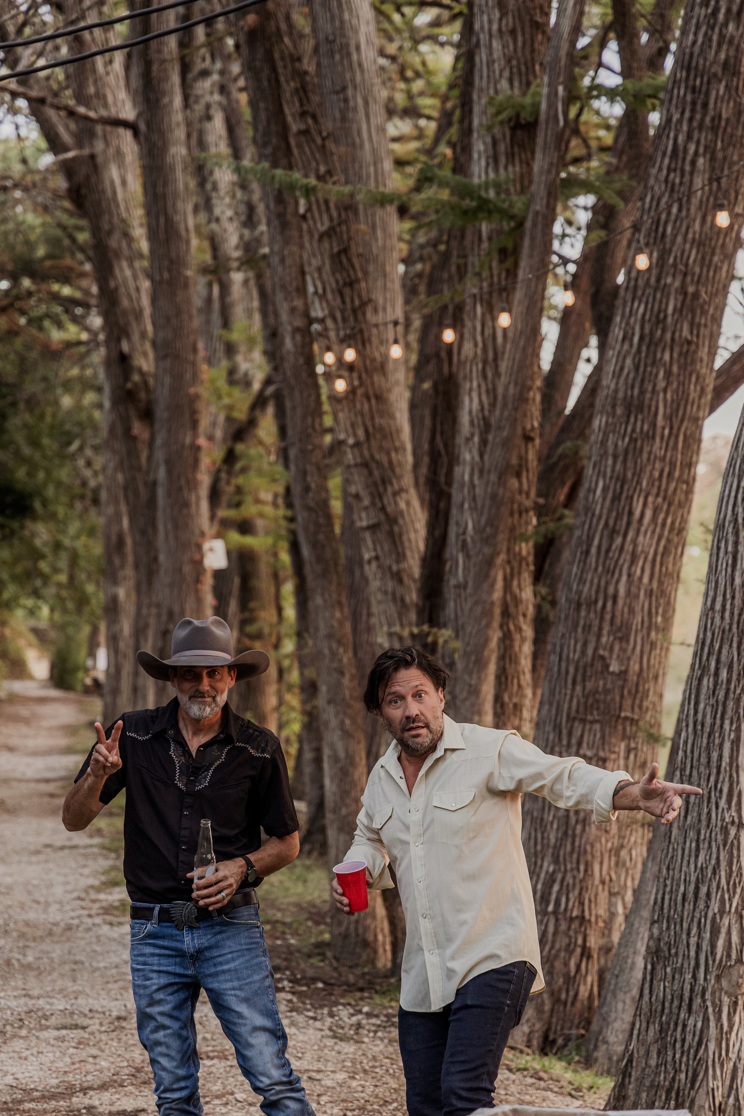 Two men at a wooded outdoor gathering, one wearing a cowboy hat and black shirt making a peace sign, the other in a light shirt pointing, with string lights hanging in the trees.