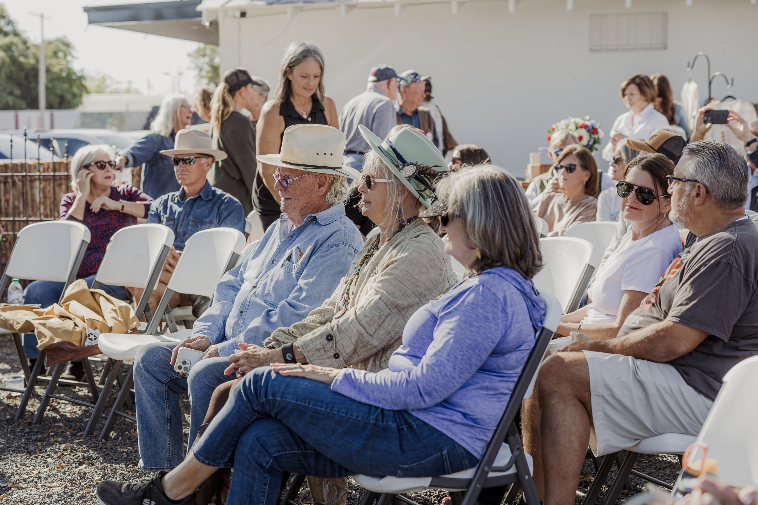 A group of people seated outdoors at an event on a sunny day, some wearing hats and sunglasses.