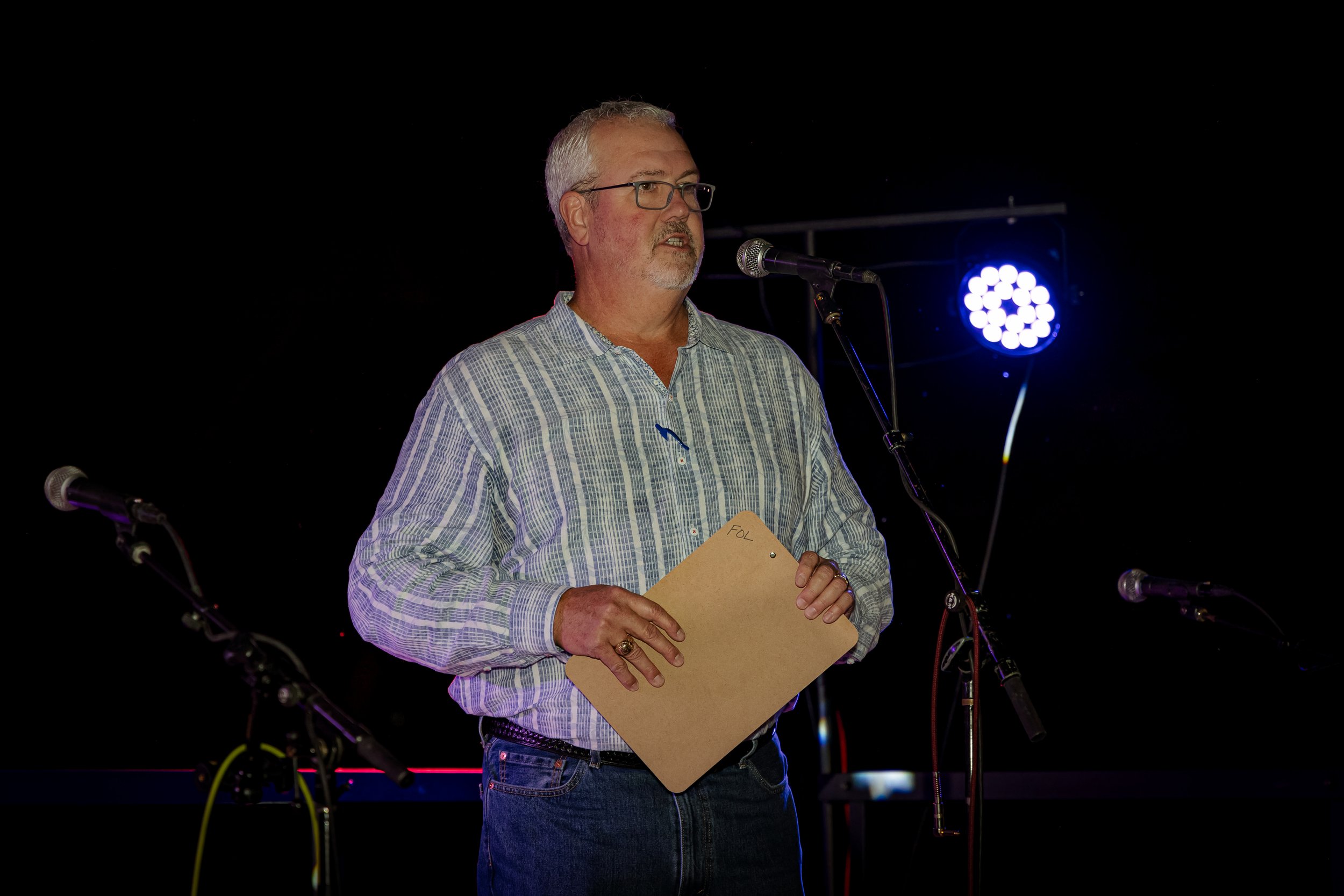 A man with glasses, gray hair, and a beard stands on stage, speaking into a microphone while holding a clipboard marked 'FOU'. He is wearing a plaid shirt and jeans, with stage lighting in the background.